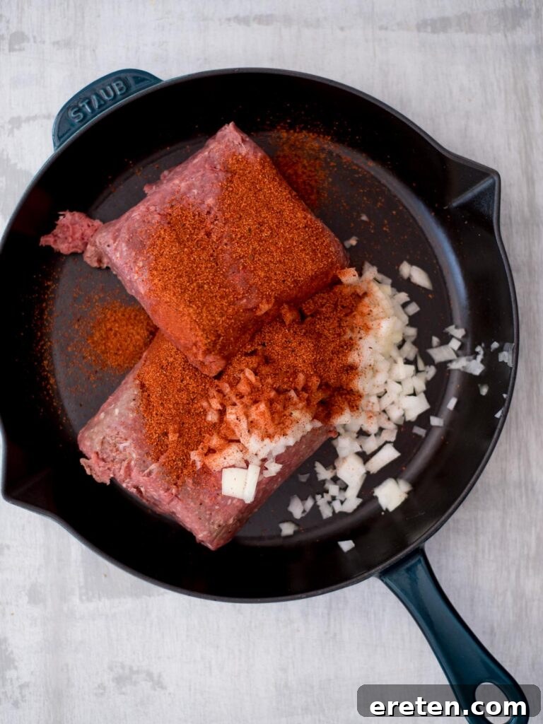 Raw ground beef, finely diced onions, and taco seasoning are added to a hot cast iron pan, ready to be cooked and combined.
