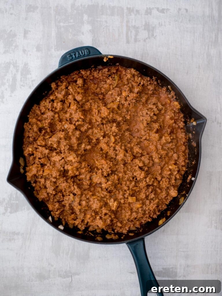 A cast iron pan filled with the perfectly seasoned ground beef mixture, now enriched with green chiles, enchilada sauce, and melted cream cheese, ready for assembly.