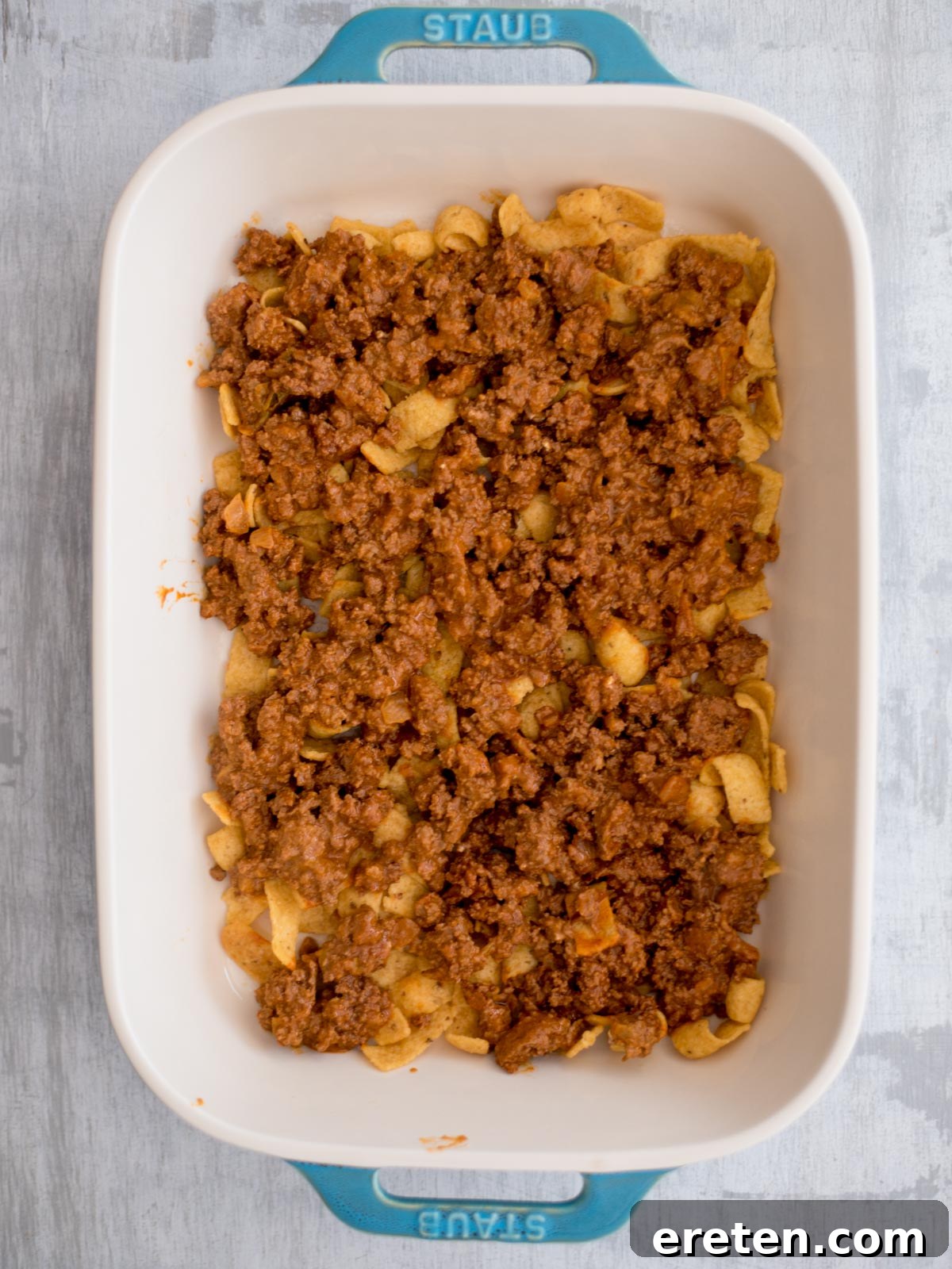 A baking dish with a base of Fritos, now topped with a rich layer of seasoned ground beef mixture, evenly spread and ready for cheese.