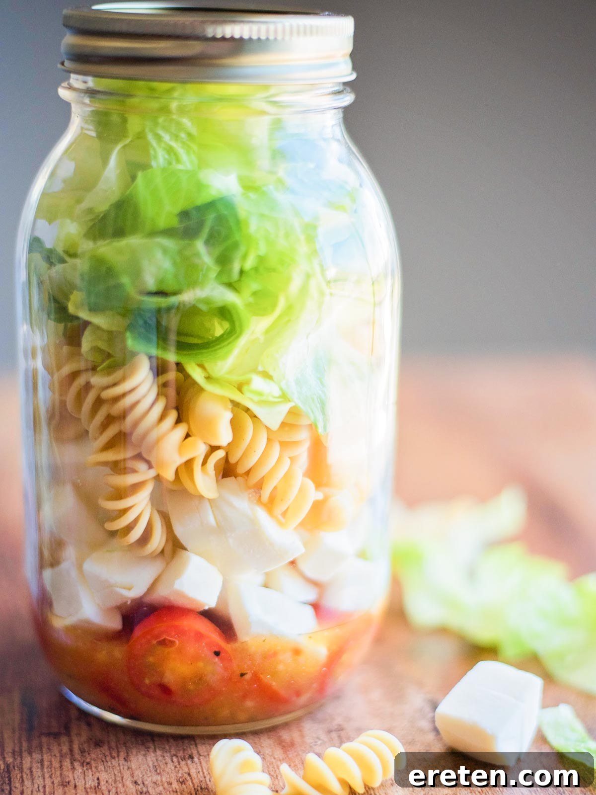 Layered Italian Jar Salads 2 Vibrant Italian Mason Jar Salad with fresh ingredients like grape tomatoes, mozzarella, pasta, and lettuce, surrounded by extra ingredients on a rustic wooden table.