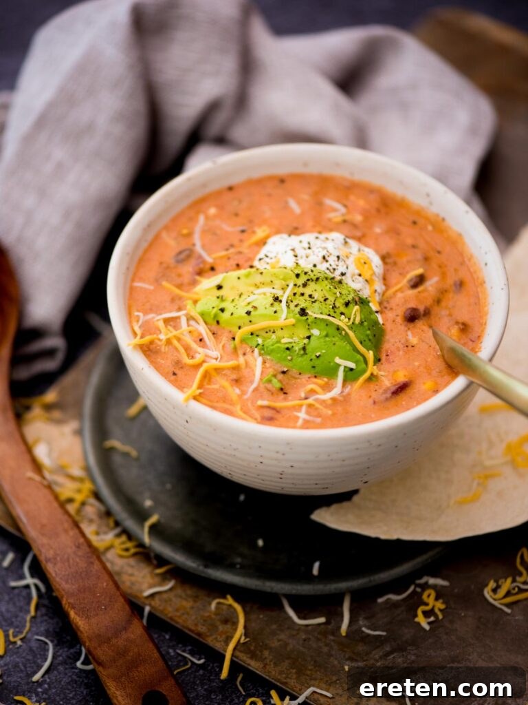 A ceramic spoon resting inside a deep bowl of creamy taco soup, garnished with swirls of sour cream and fresh avocado slices.