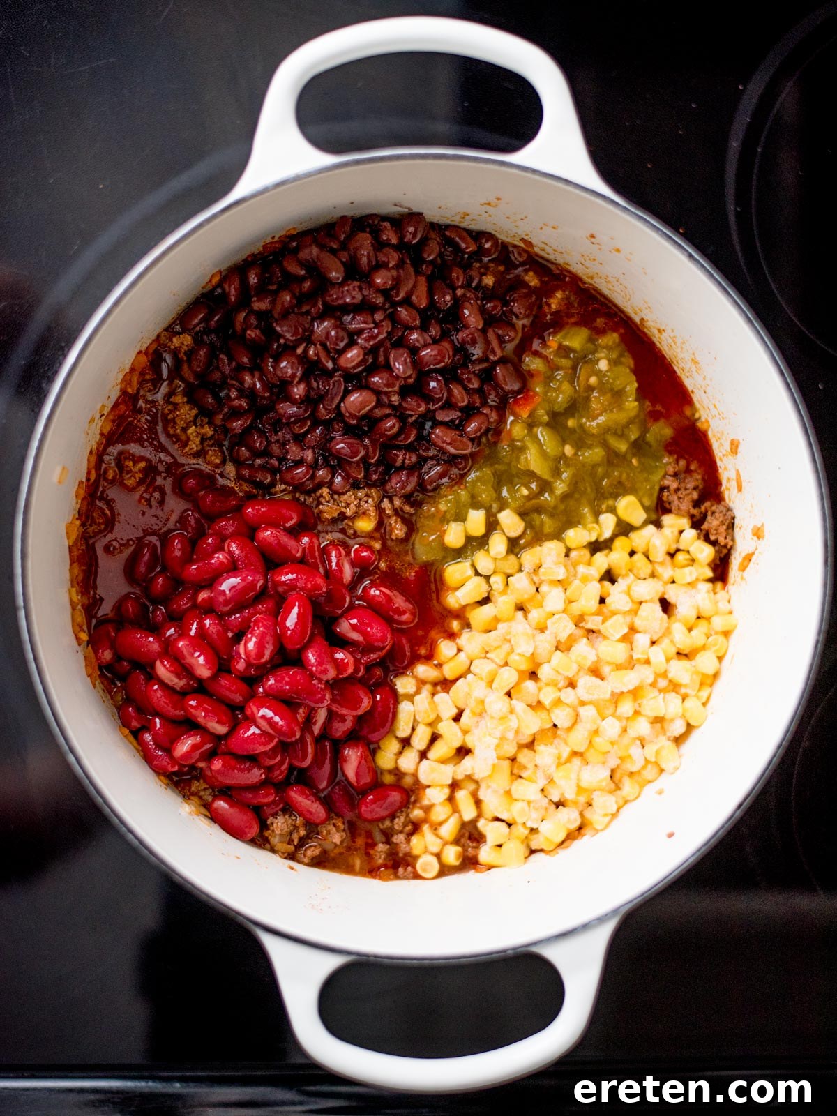 A dutch oven containing cooked ground beef and onions, with corn, black beans, and kidney beans newly added.