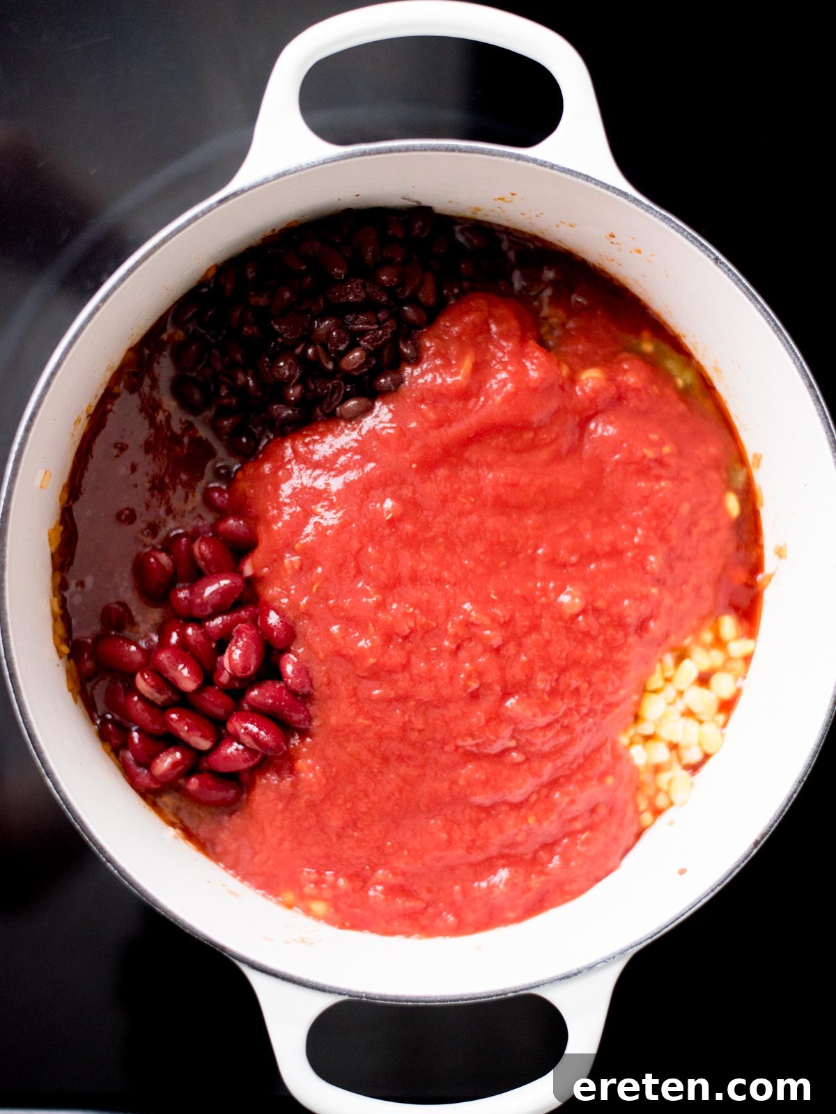 Crushed tomatoes being poured over the mixture of ground beef, onions, and beans in a dutch oven.