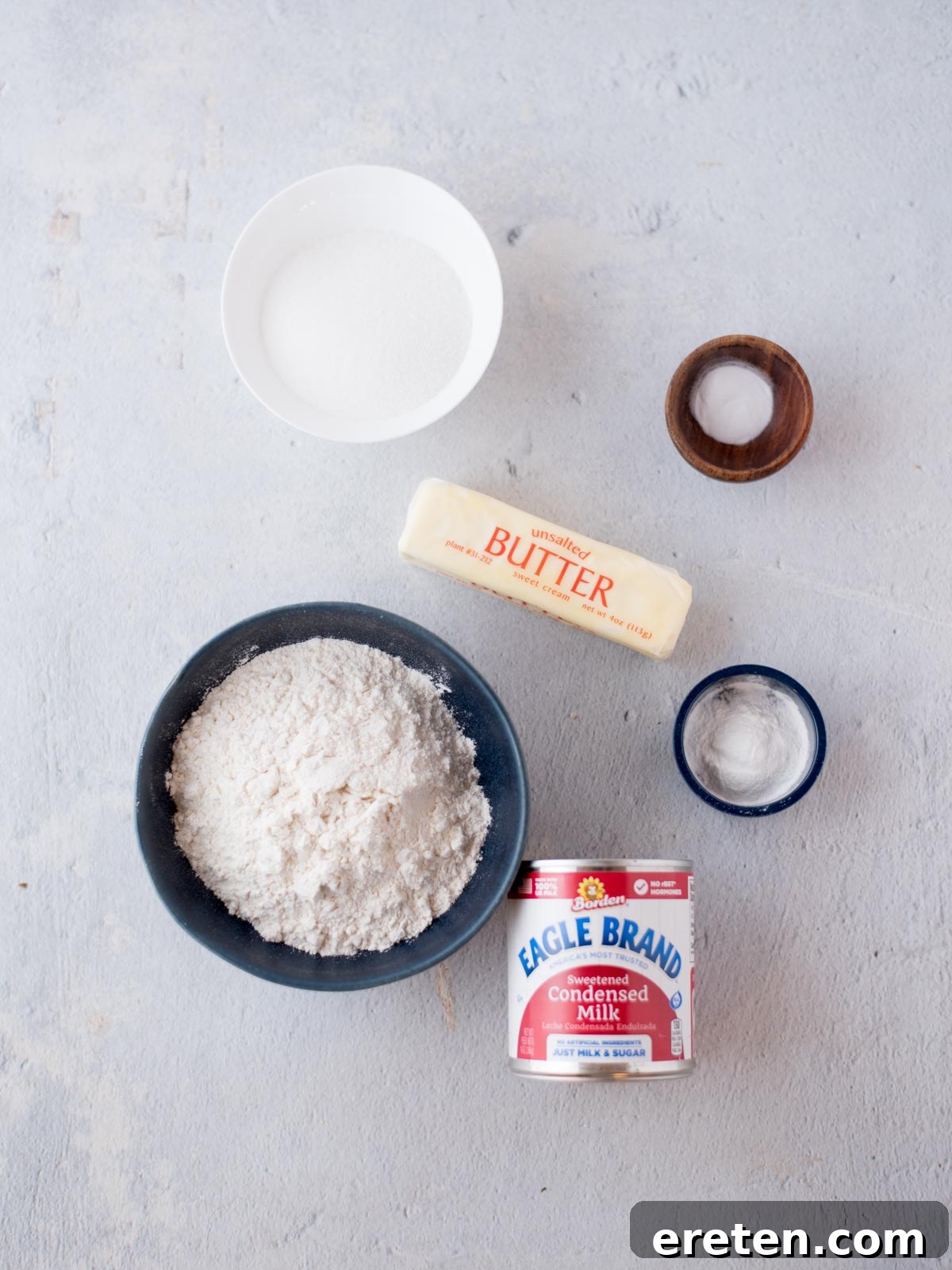 Individual ingredients for condensed milk cookies laid out on a white surface, including flour, butter, sugar, and a can of condensed milk