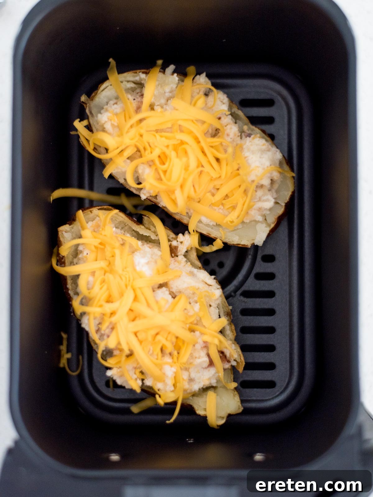 Two twice baked potatoes topped with shredded cheese in the air fryer ready to cook.