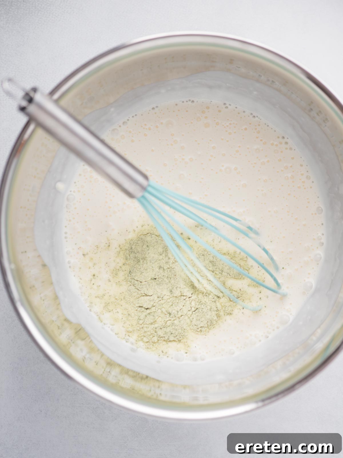 A dry ranch seasoning packet being added to the creamy buttermilk and mayonnaise mixture in a mixing bowl.