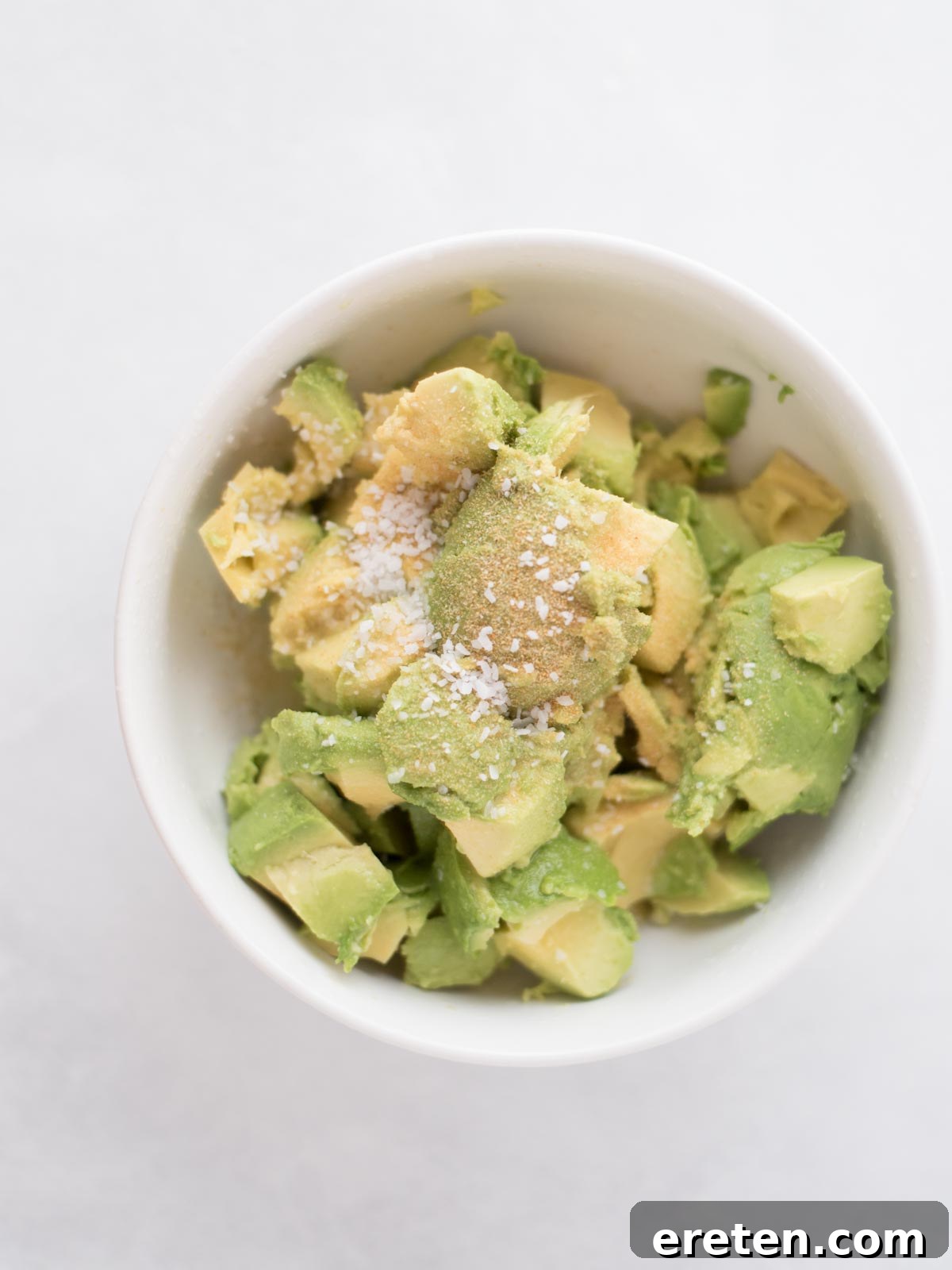 Chopped avocado in a bowl, generously seasoned with garlic powder and coarse salt, awaiting the addition of lime juice and zest.