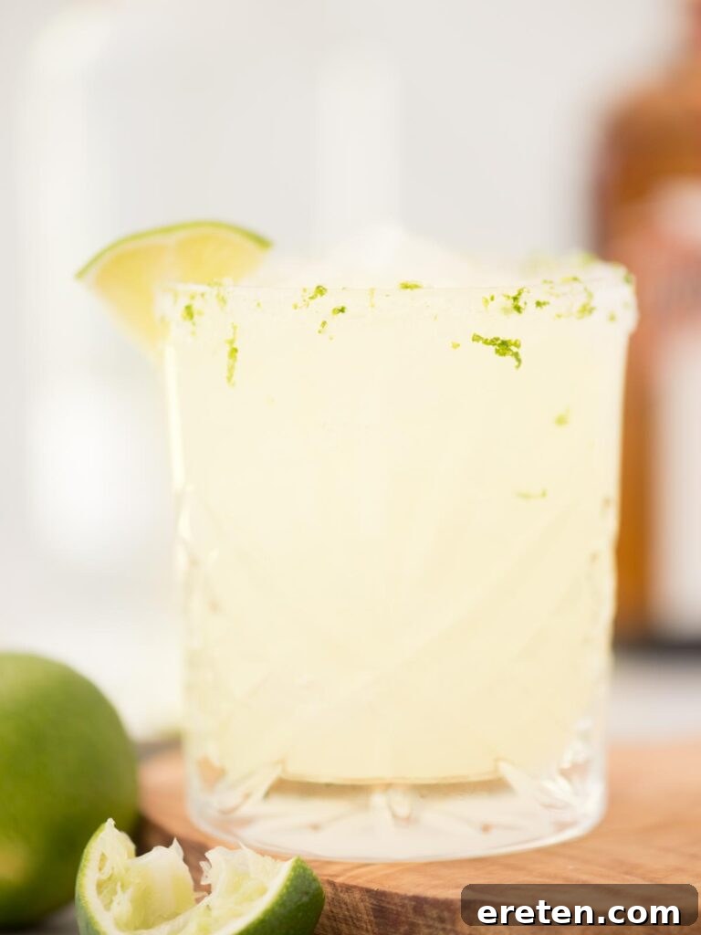 Stone Cold Margarita 2 margarita on the rocks in a salt and lime zest rimmed glass and a lime wedge sitting on a wooden board with an extra lime to the side