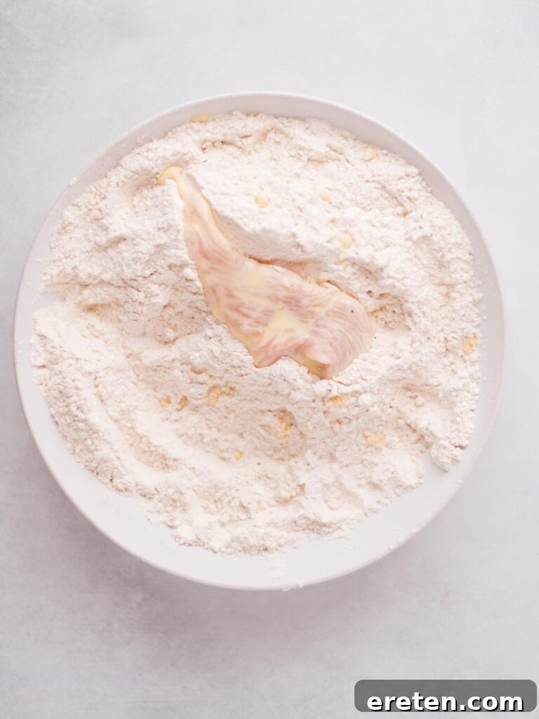 Chicken piece being dipped in egg mixture, then transferred to a bowl of seasoned flour for coating, illustrating the double-breading technique.