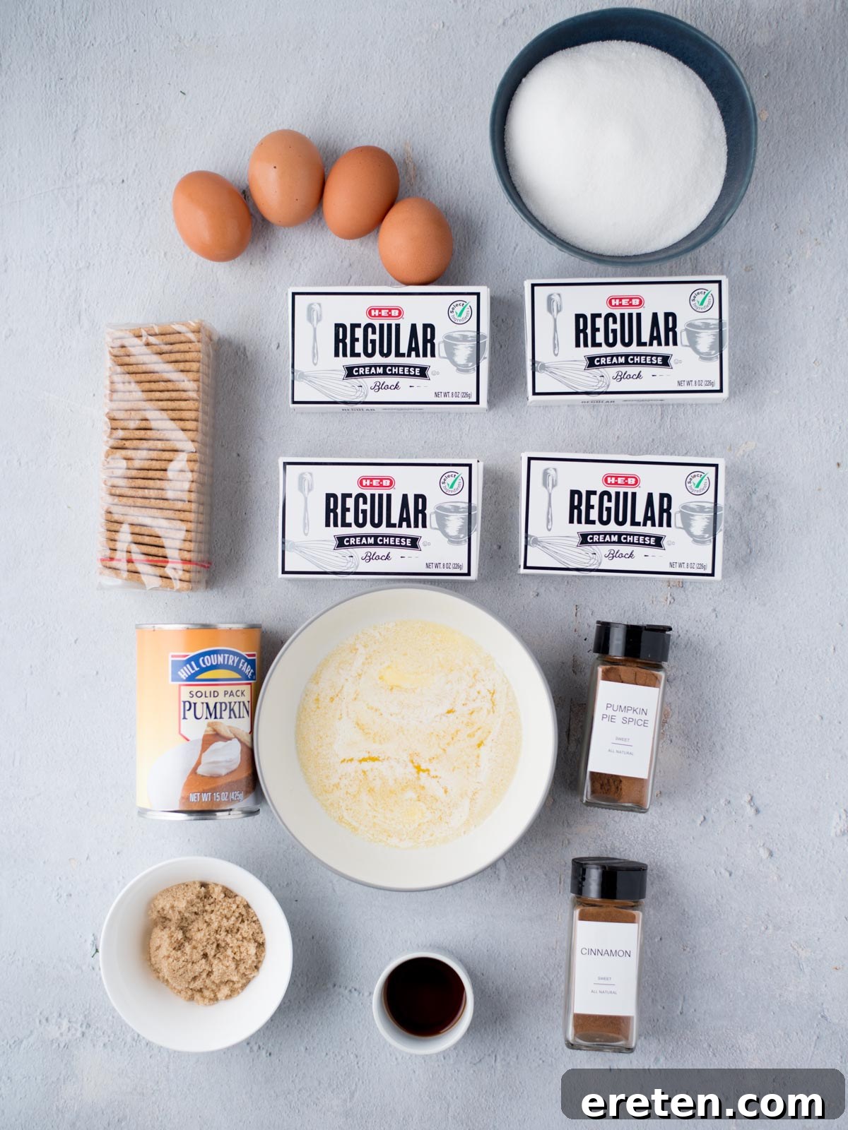 A collection of fresh ingredients laid out on a surface, ready to be used for making pumpkin cheesecake bars, including cream cheese, pumpkin puree, eggs, spices, and graham crackers.