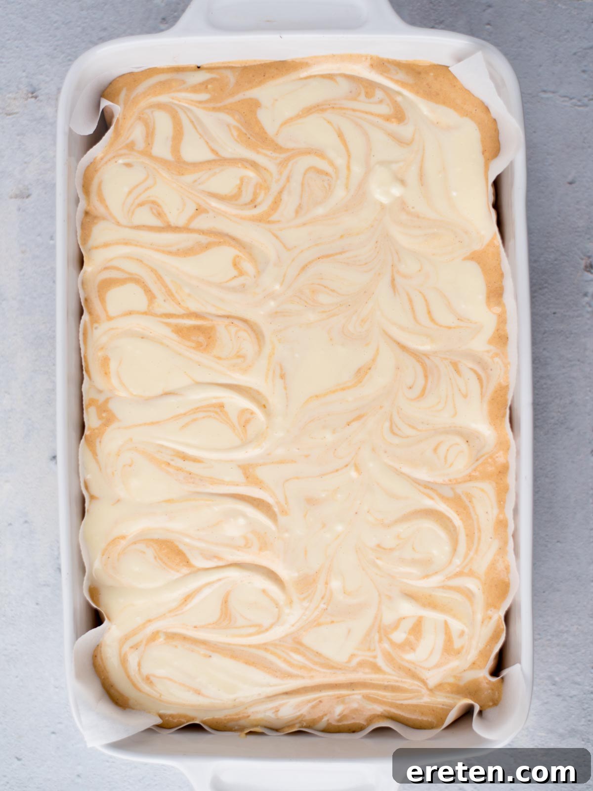 Unbaked pumpkin cheesecake bars with elegant swirls of plain cheesecake batter over the pumpkin layer in a 9x13 inch dish.