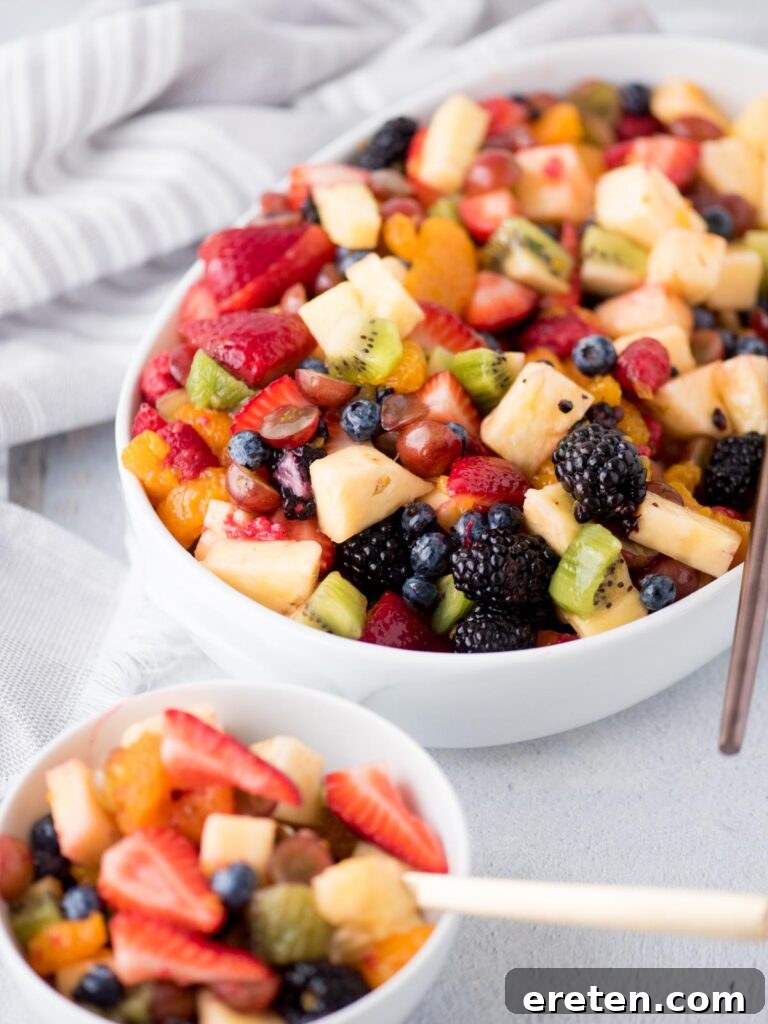 A generous portion of fruit salad presented in a white bowl with a serving spoon, alongside a smaller, individual serving in a separate white bowl with a fork, ready for enjoyment.