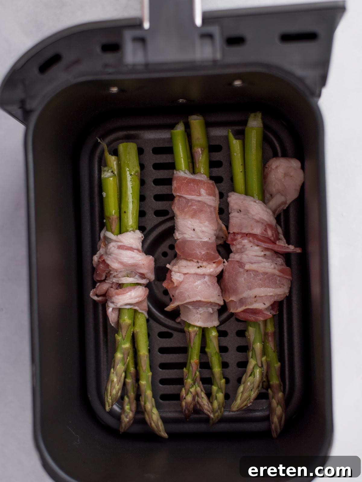 Crispy Air Fryer Bacon Asparagus Bundles 4 Seasoned asparagus wrapped in bacon and laying in an air fryer basket.