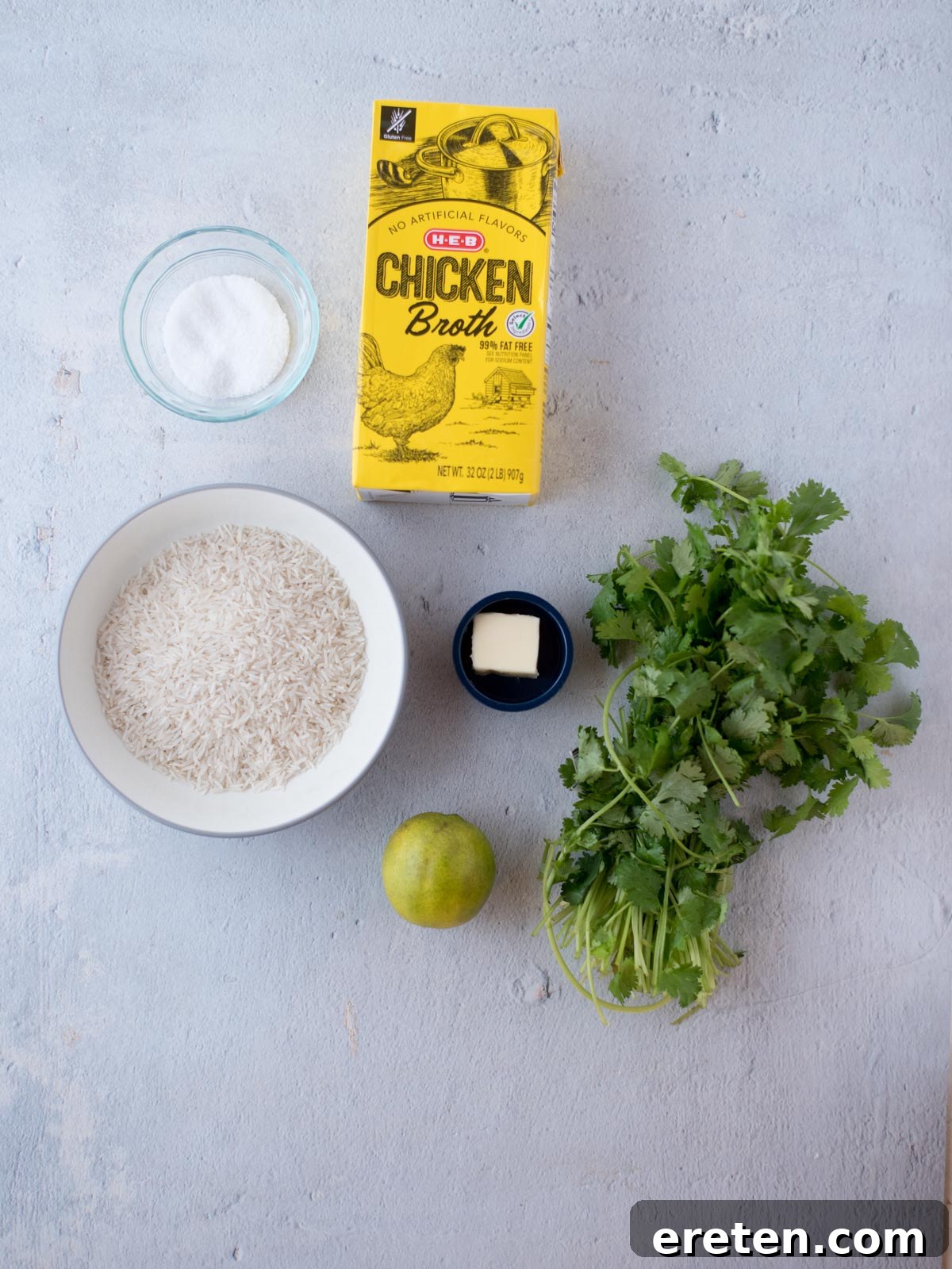 All the fresh ingredients laid out on a wooden surface: fresh cilantro, whole limes, a bag of rice, a stick of butter, and a container of chicken stock, ready for cooking.