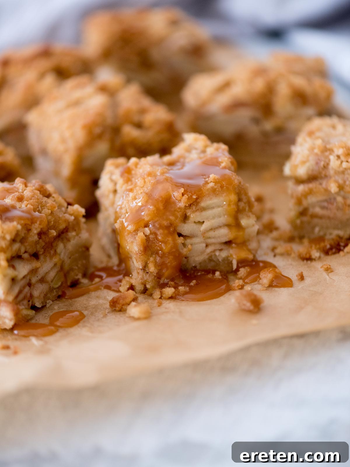 Close-up of apple crumble bars with layers of apple slices and a crumbly topping. Drizzled with caramel sauce, the bars sit on a piece of parchment paper, creating a delicious and inviting dessert scene.