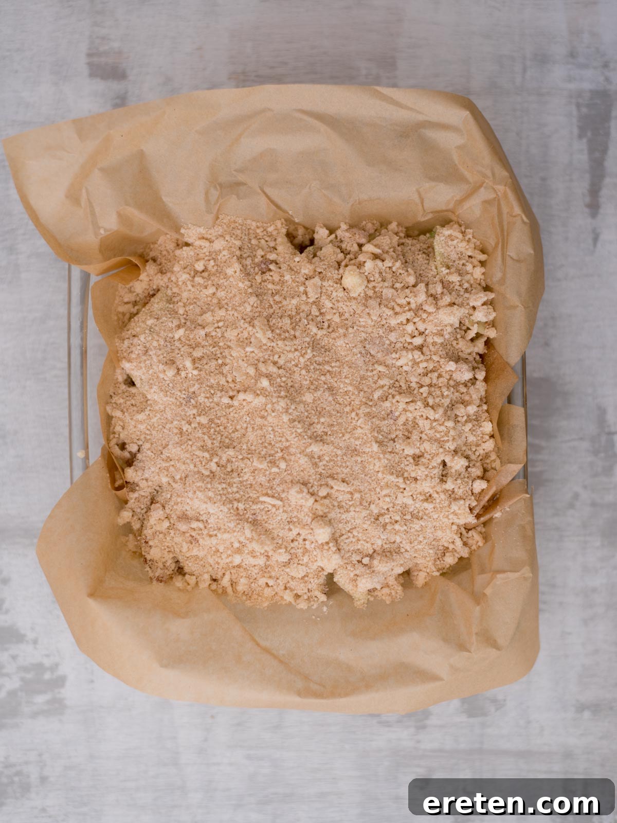 A baking dish lined with brown parchment paper is filled with an unbaked, crumbly, cinnamon-colored topping, ready to be baked. The background is a light gray surface.