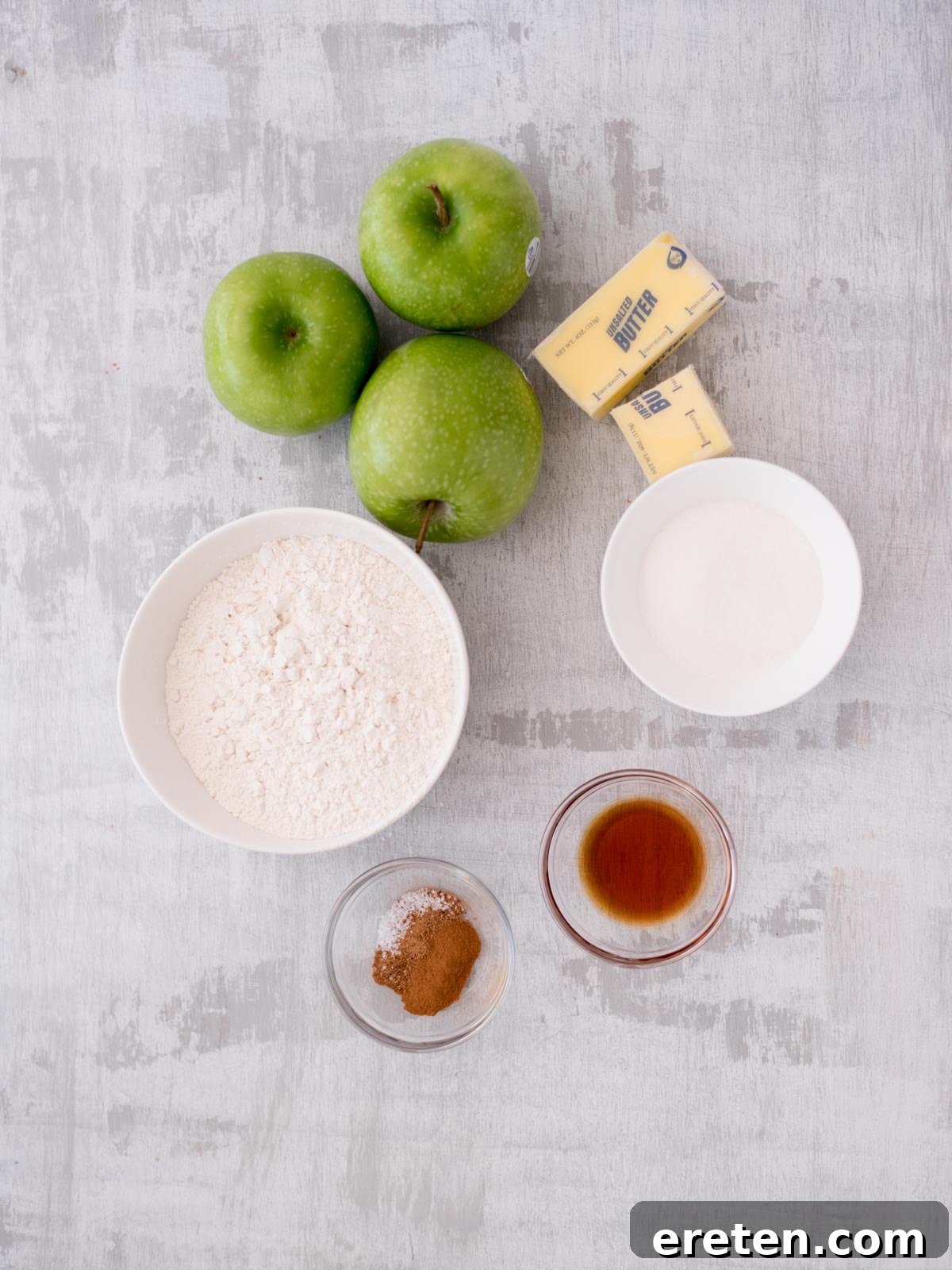 Top-down view of baking ingredients on a gray surface, including three green apples, two sticks of butter, a bowl of flour, a bowl of sugar, and small bowls with spices and vanilla extract.
