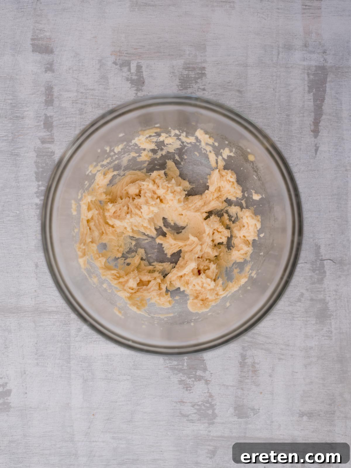 A glass bowl containing creamy, partially mixed batter is placed on a light gray wooden surface. The batter is unevenly blended, with some visible lumps.