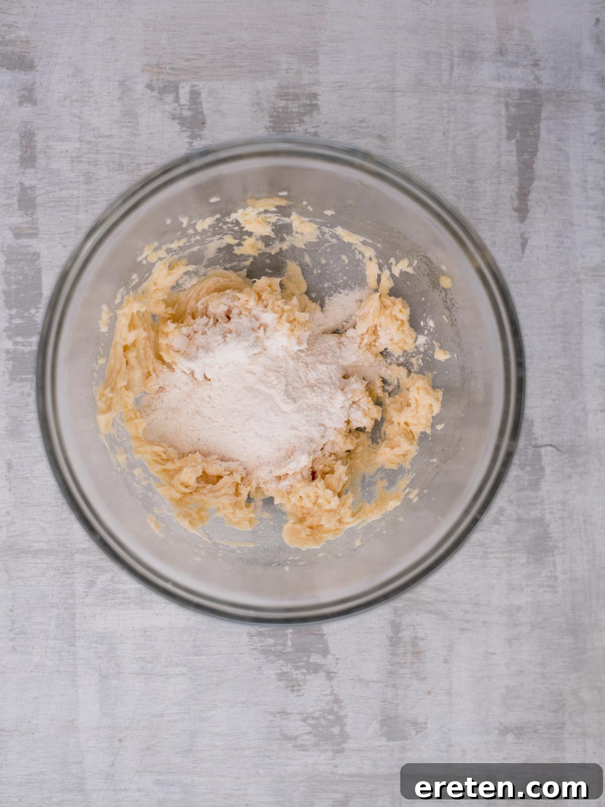 A glass bowl contains partially mixed dough with flour and softened butter on a textured gray surface. The ingredients are in the early stages of forming a cohesive dough mixture.