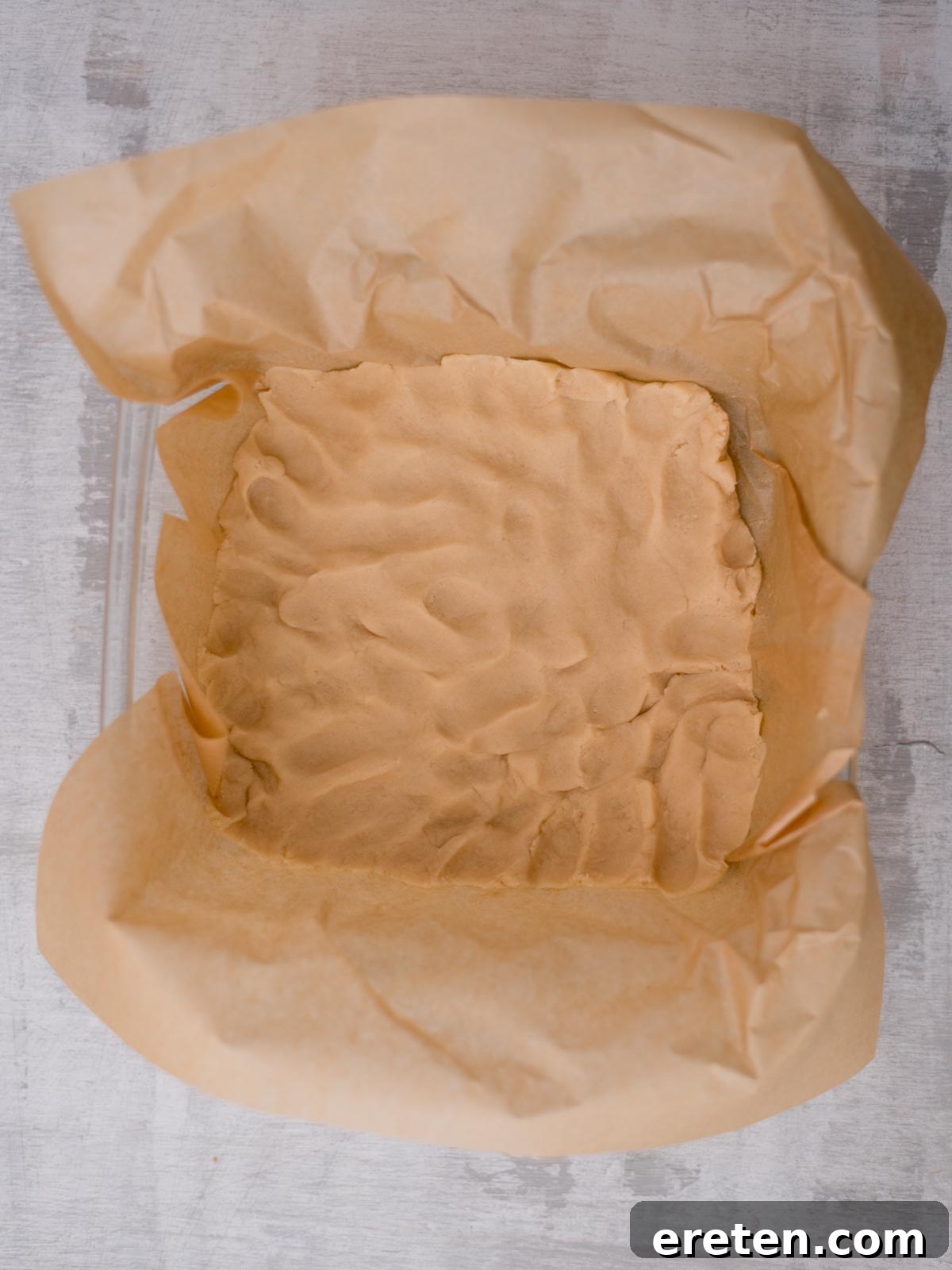 A square baking pan lined with parchment paper containing an evenly pressed layer of raw shortbread dough. The dough has a slightly textured surface. The pan is placed on a gray countertop.