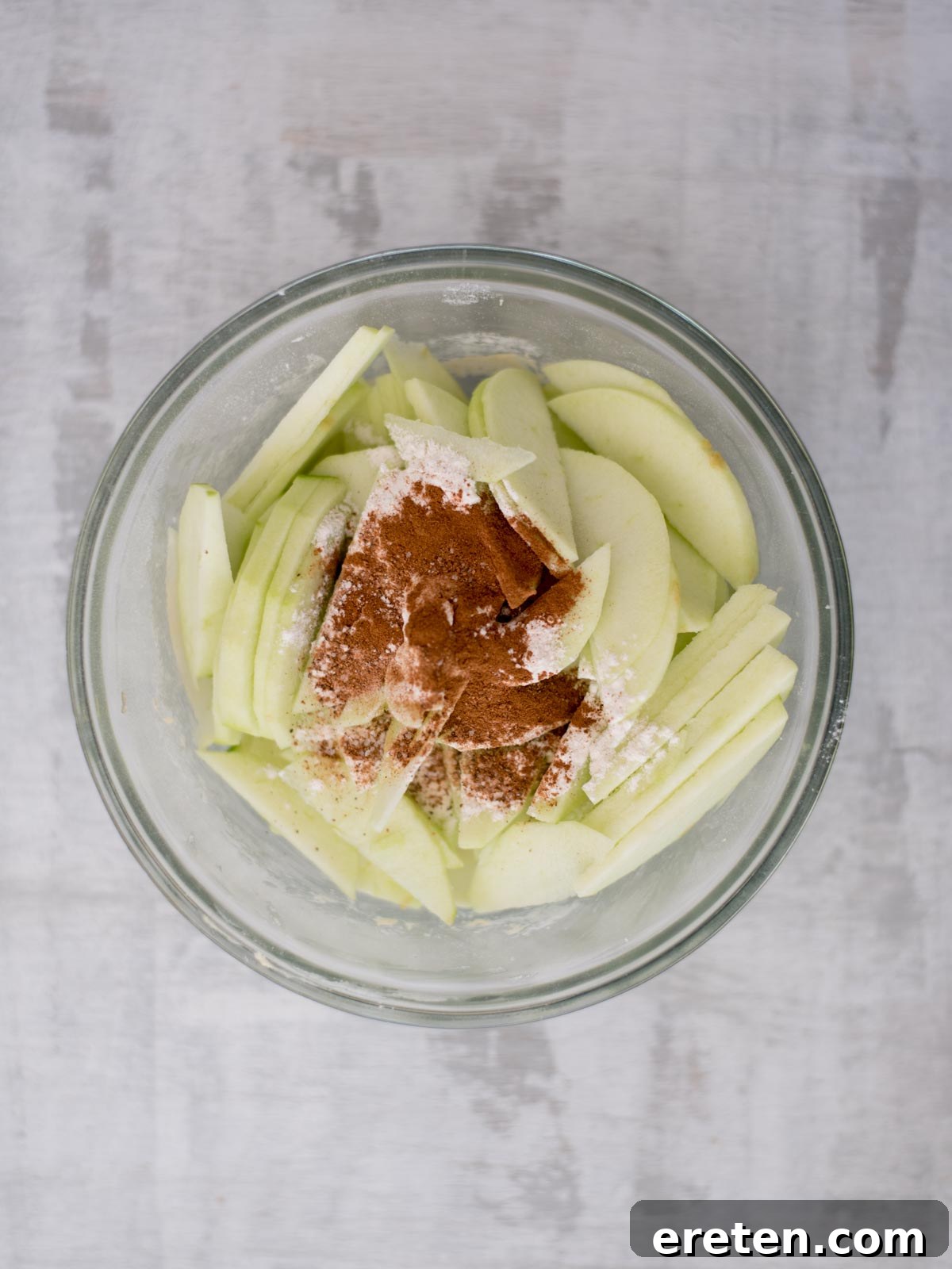 A glass bowl containing sliced green apples, sprinkled with cinnamon and flour, placed on a light gray surface.