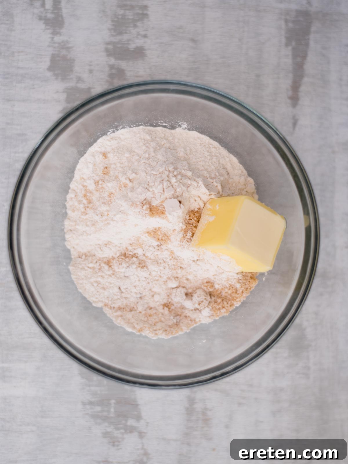 A glass bowl contains baking flour and a block of butter, ready for mixing, placed on a gray surface.
