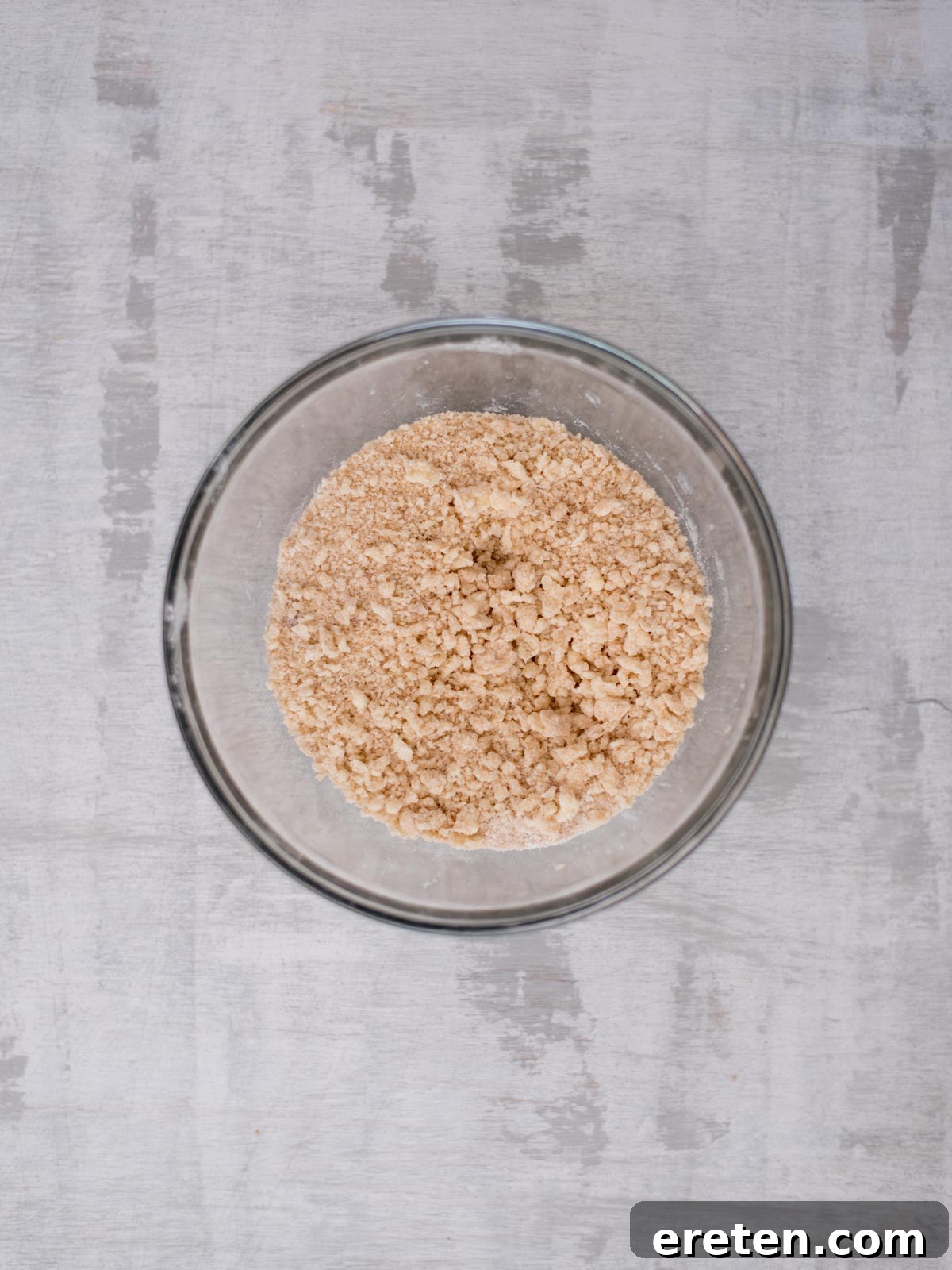 A glass bowl filled with a crumbly mixture of light brown, dry ingredients sits on a textured gray surface, viewed from above.