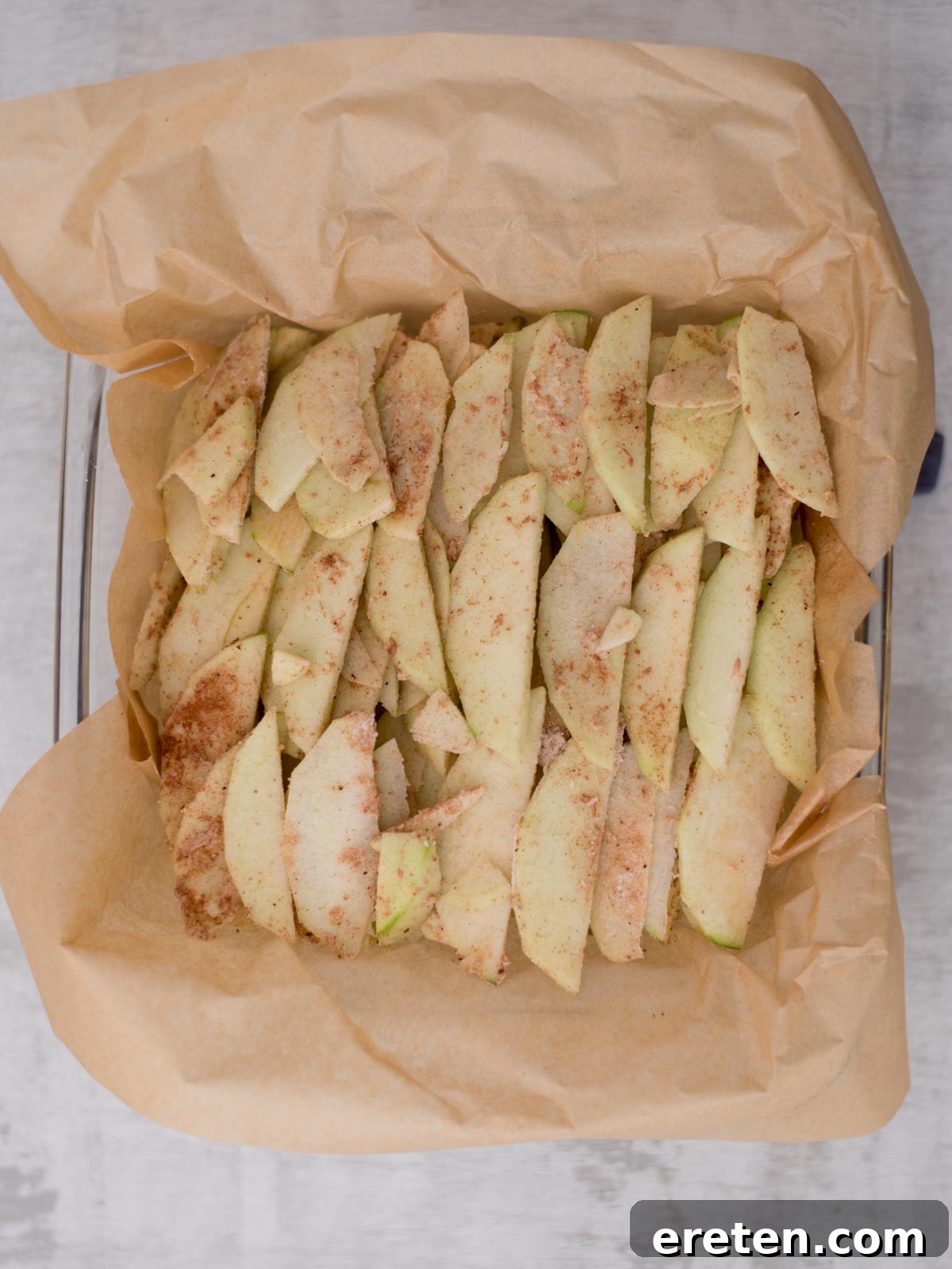 Sliced apples seasoned with cinnamon on parchment paper in a baking dish.