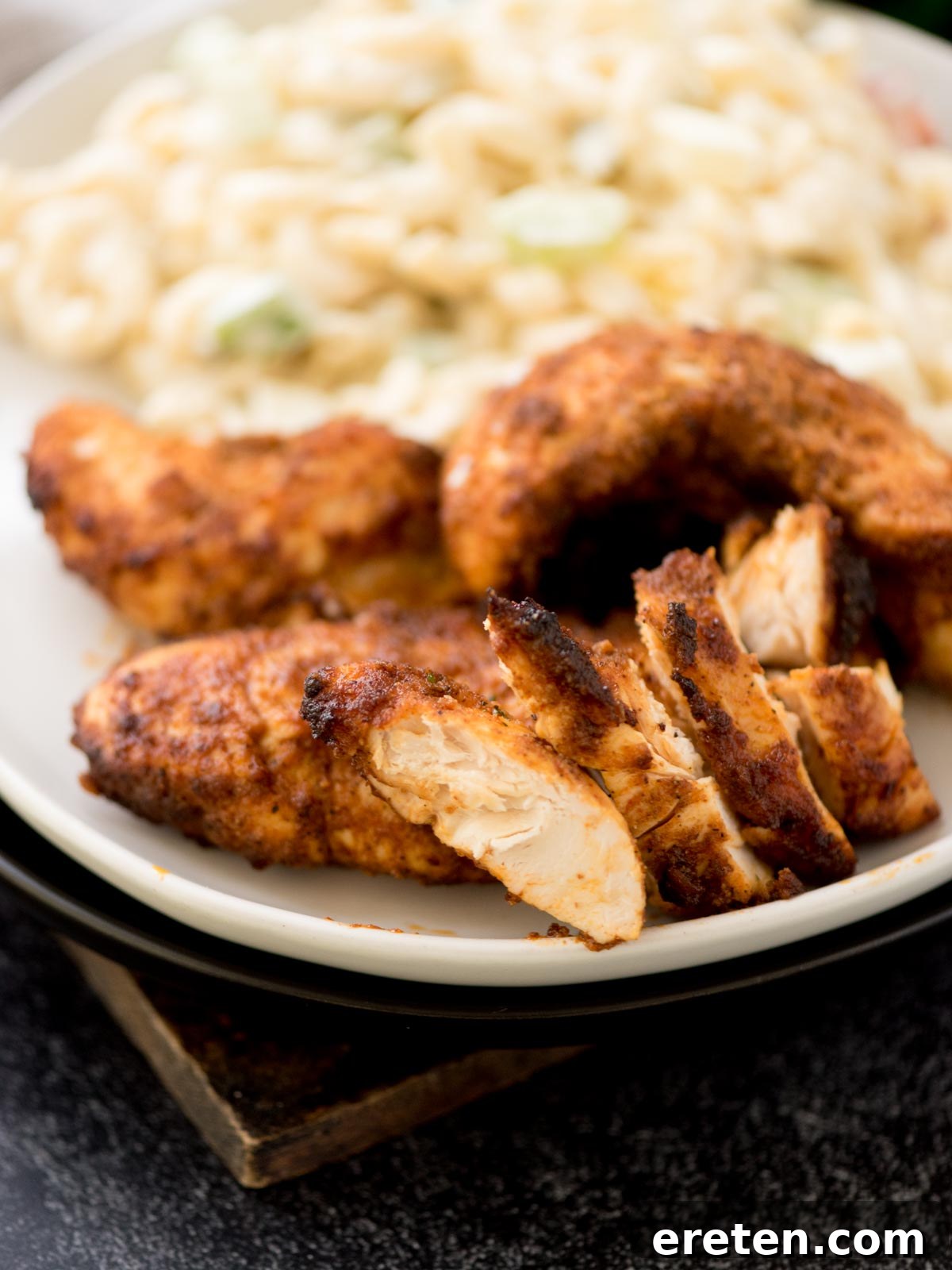 Golden-brown air fryer chicken tenders resting on a white plate, accompanied by a creamy pasta salad, all beautifully arranged on a dark table surface.