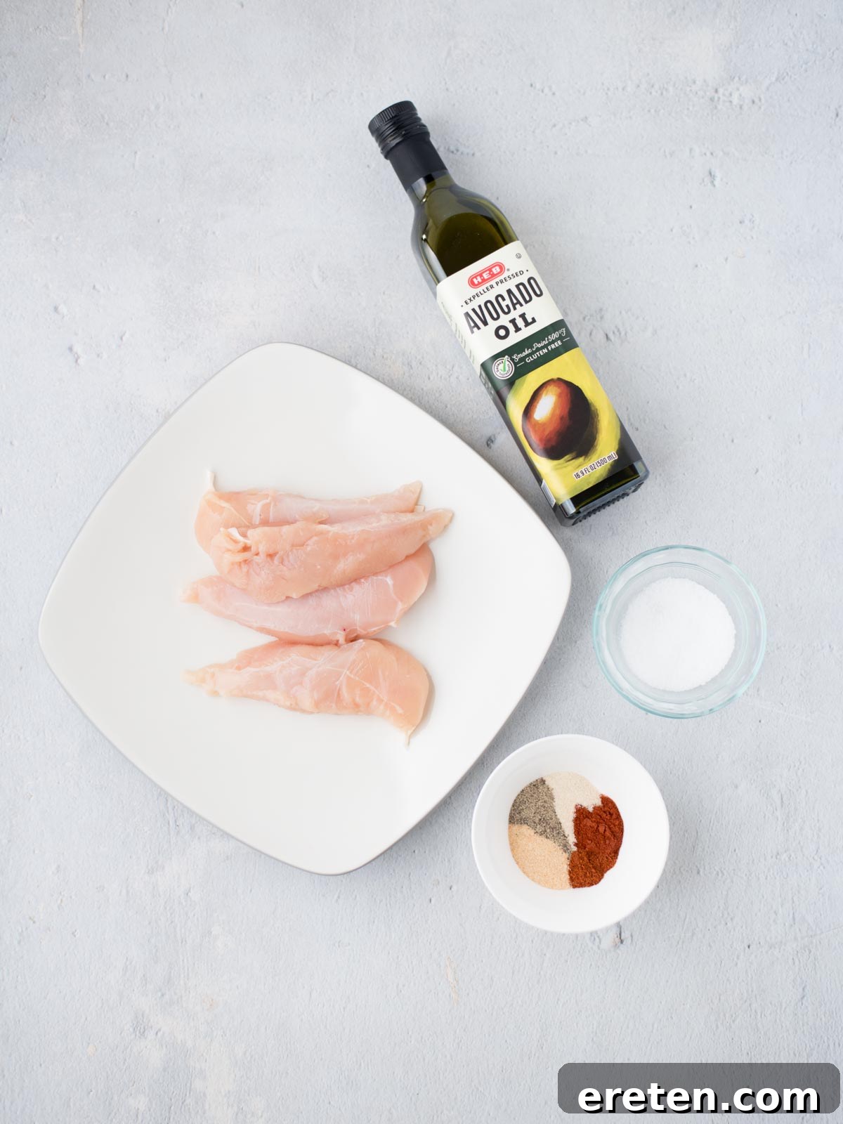 Raw chicken tenderloins on a square white plate, accompanied by a bottle of avocado oil and small bowls of various spices including paprika, black pepper, and garlic powder, all arranged on a light gray surface, ready for seasoning.