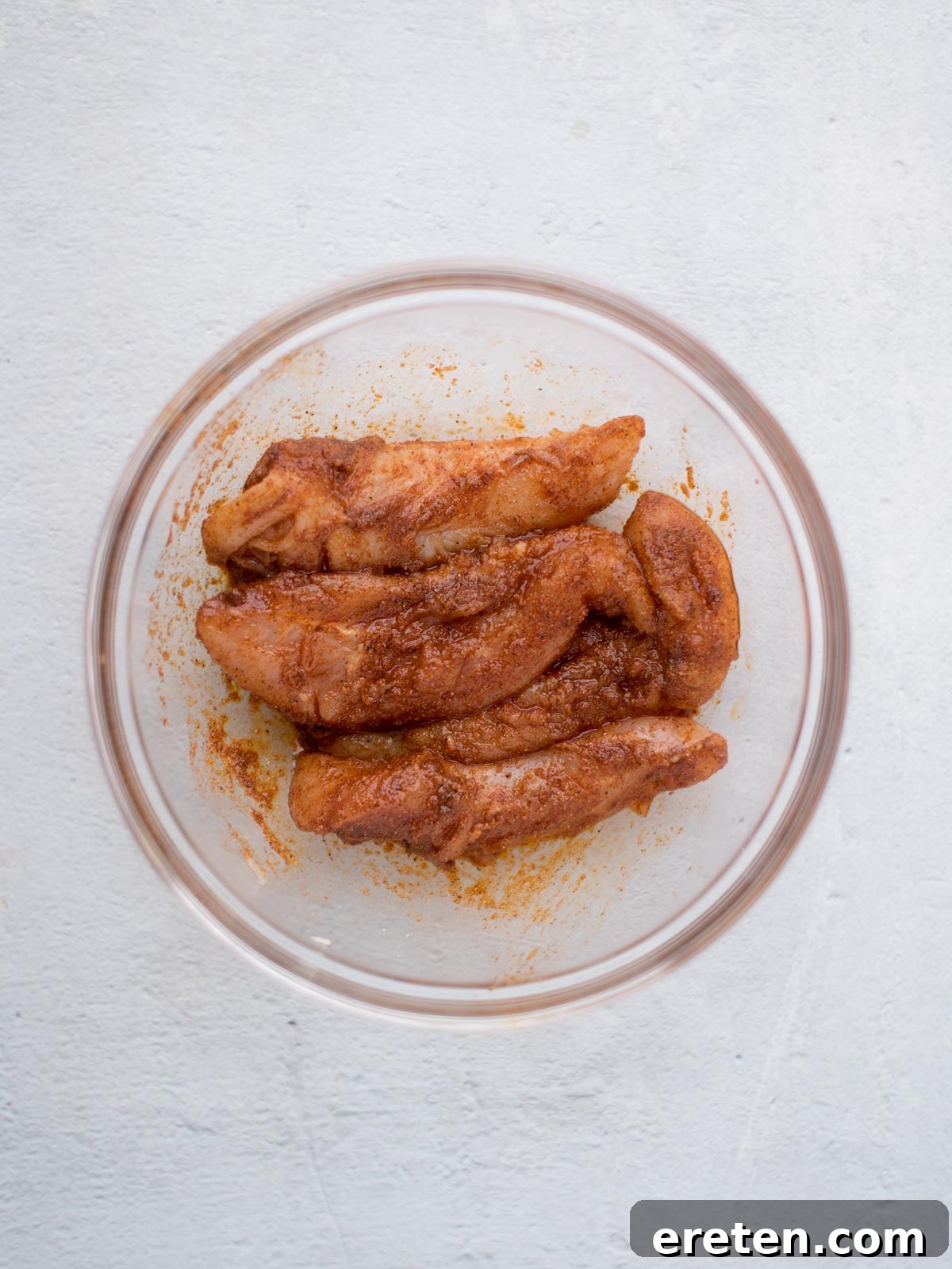 Raw chicken tenderloins coated generously with a vibrant spice blend, resting in a clear glass bowl on a light gray surface, ready for air frying.