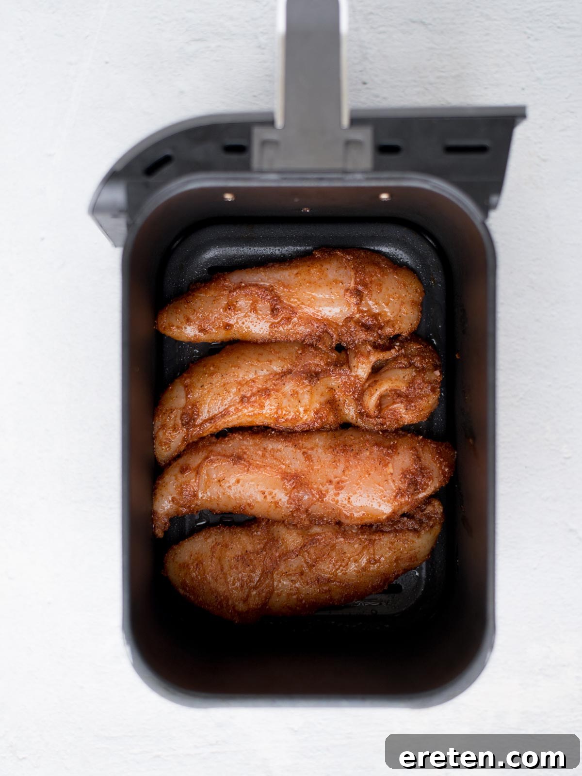 Four golden-brown, seasoned air fryer chicken tenders arranged neatly in an air fryer basket, captured from a top-down perspective on a light surface, indicating they are perfectly cooked or ready for the next step.