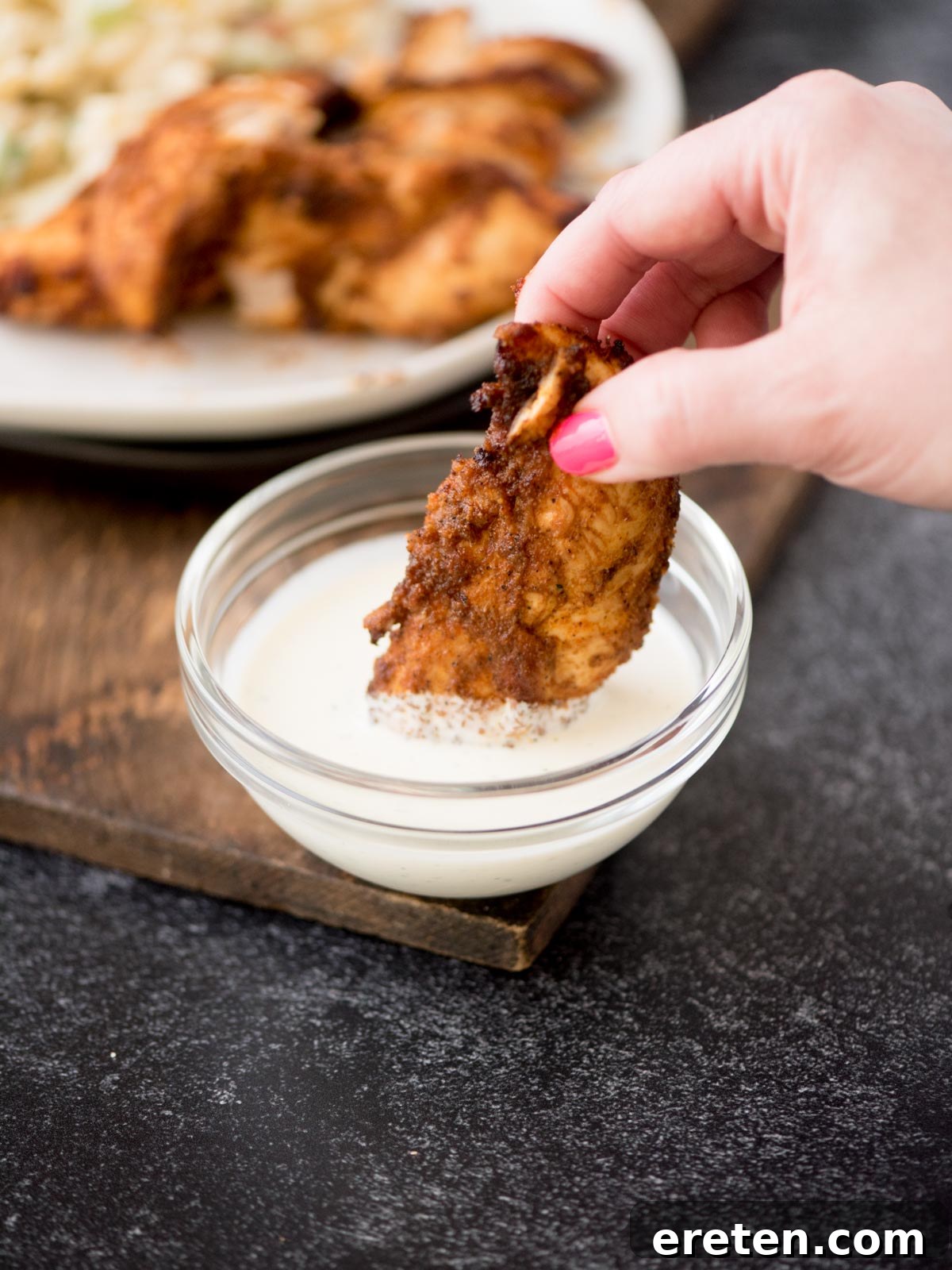 A hand with pink nail polish is seen dipping a piece of cooked, seasoned air fryer chicken tender into a small, clear glass bowl of white creamy sauce, set against a dark background with more chicken tenders on a white plate in the blurred background.