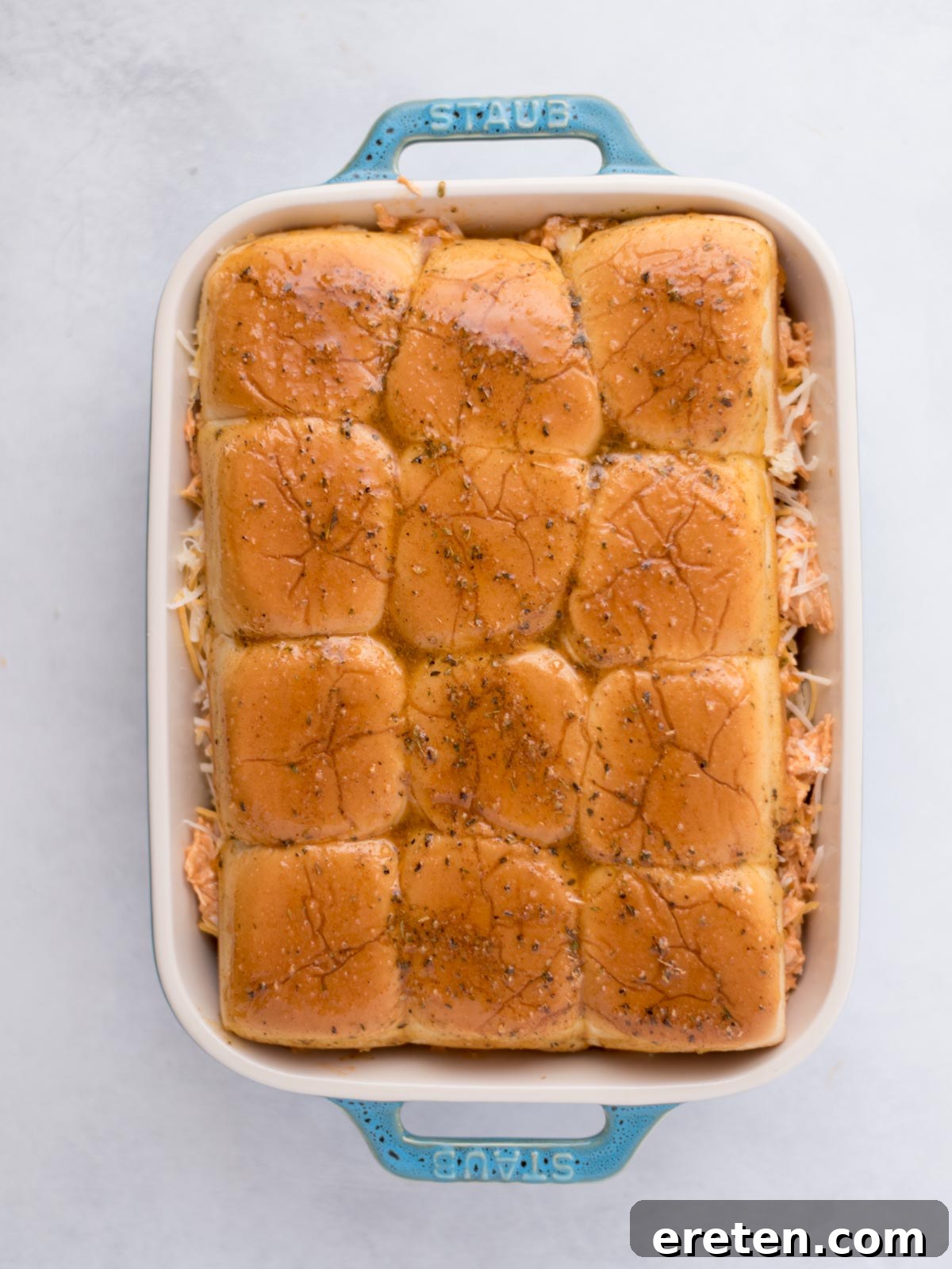 Seasoned melted butter poured over buffalo chicken sliders in a baking dish before baking.