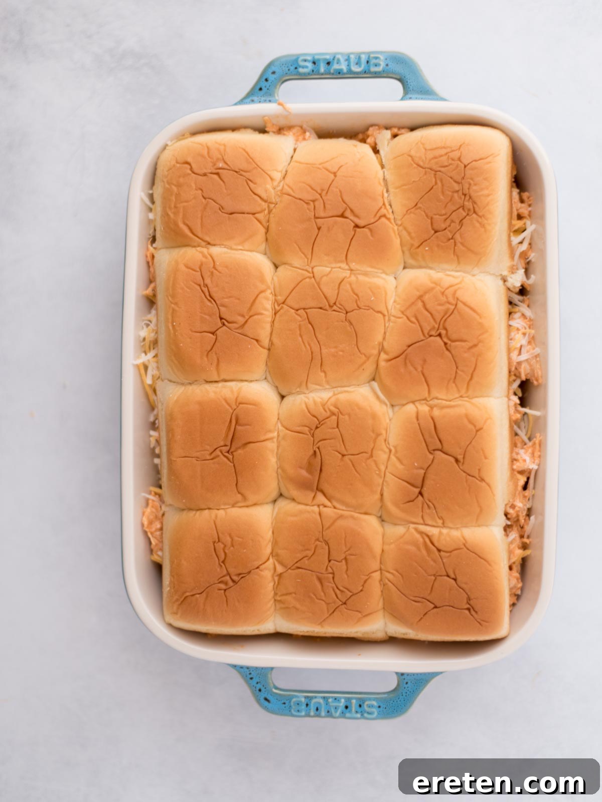 Sliders rolls topped with seasoned meat and shredded cheese in a baking dish.