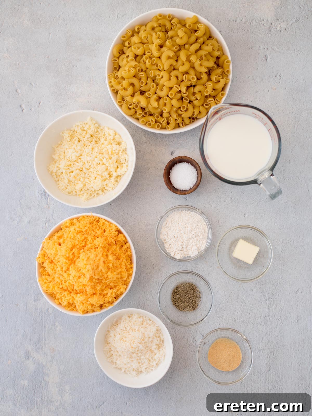 Various essential ingredients for baking mac and cheese are neatly arranged on a rustic wooden table, including uncooked elbow macaroni, blocks of sharp cheddar, Gruyere, and Parmesan cheese, fresh unsalted butter, all-purpose flour, a bottle of whole milk, and small bowls of garlic powder, coarse salt, and ground black pepper. The setup emphasizes fresh, high-quality components.
