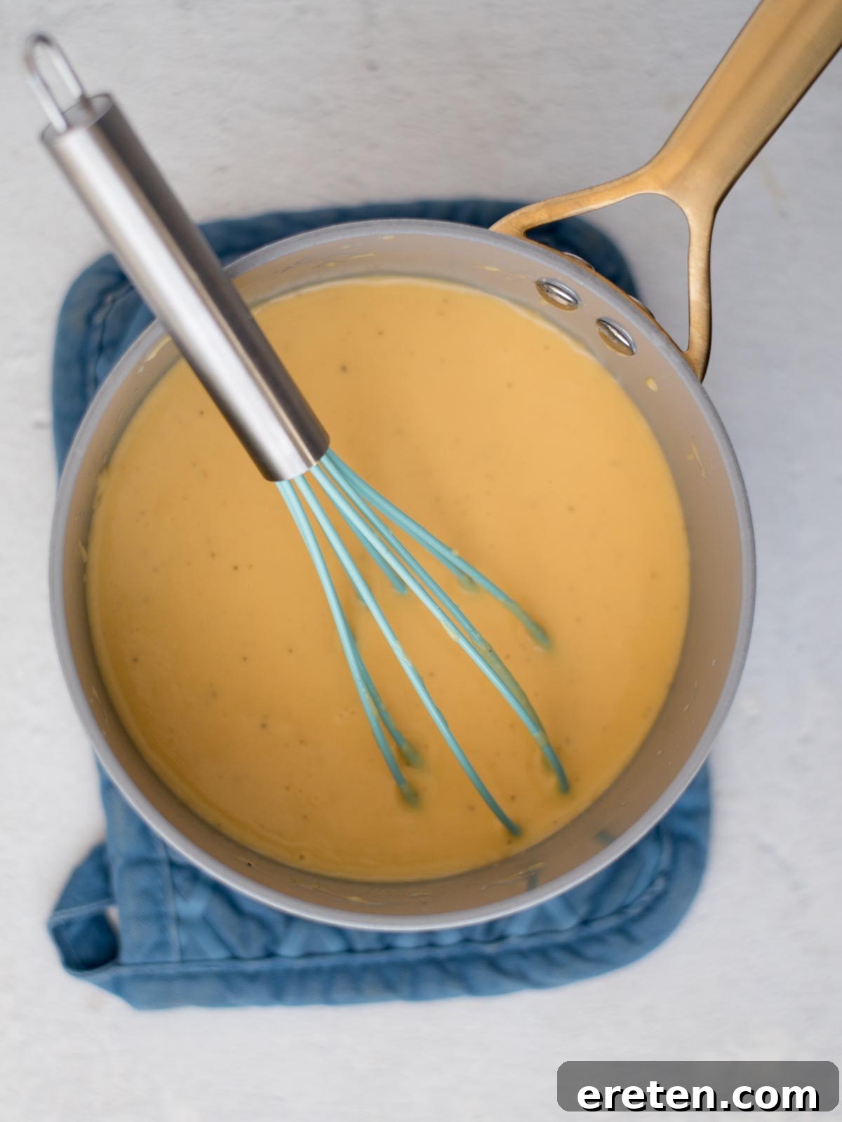A whisk resting in a saucepan of smooth, creamy, and perfectly blended cheese sauce, indicating readiness to be combined with cooked pasta.