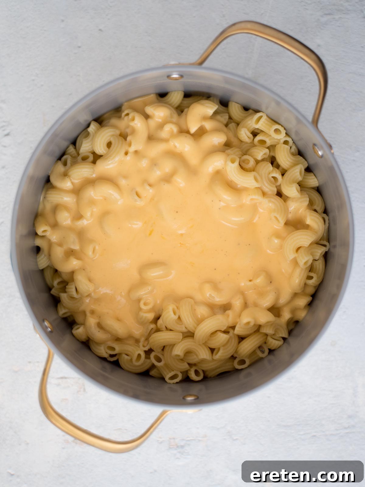 Rich, creamy cheese sauce being generously poured over cooked pasta in a saucepan, preparing for thorough mixing to coat every noodle.
