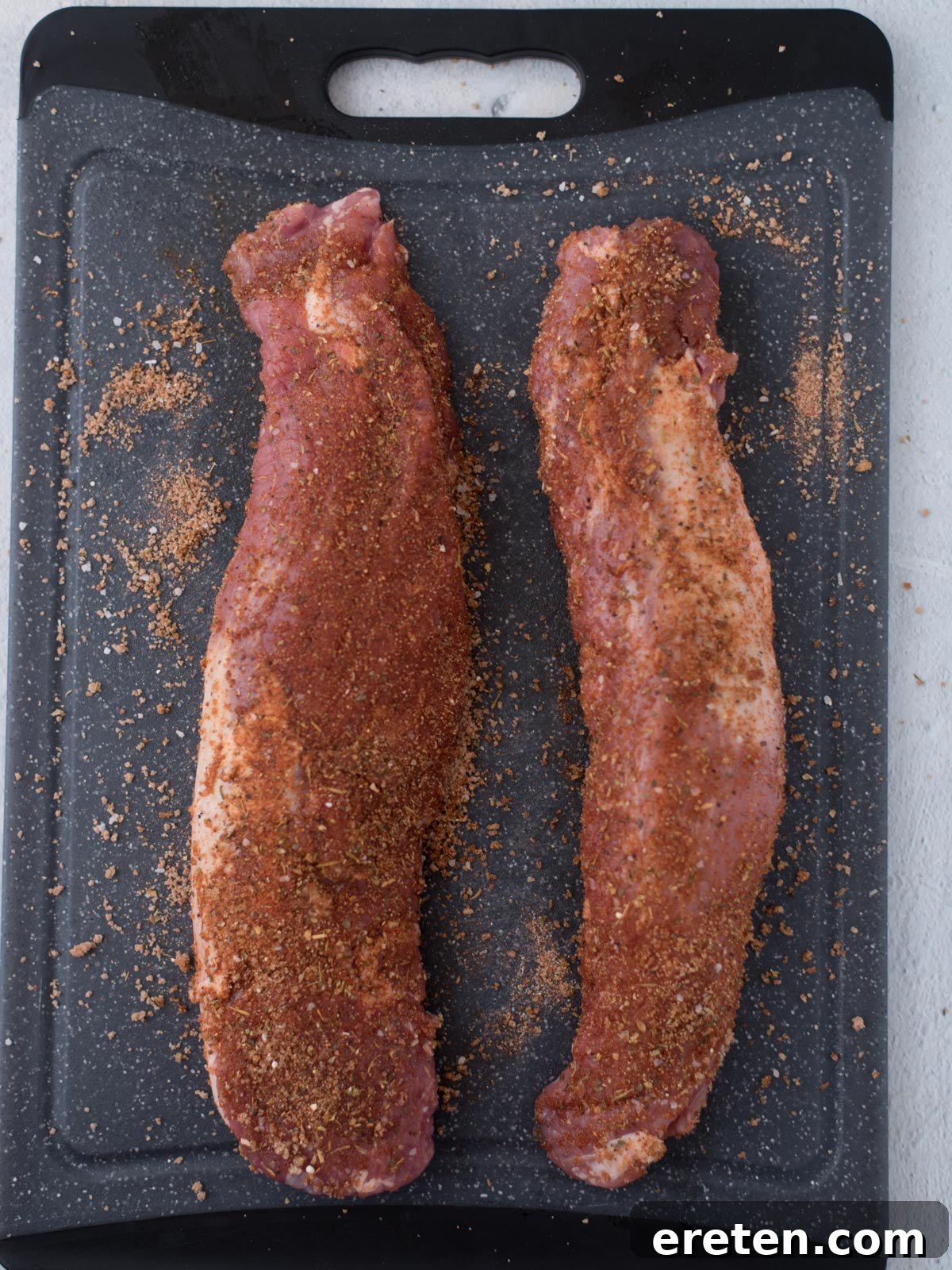 A raw pork tenderloin generously coated with a savory spice rub, resting on a black cutting board before baking.