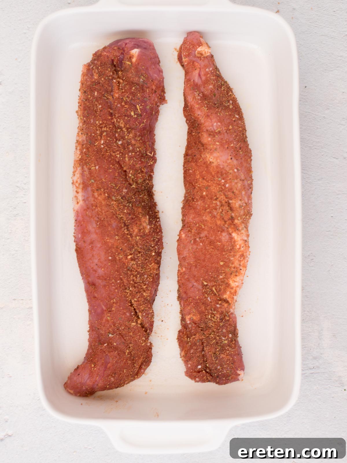 The seasoned raw pork tenderloin is carefully placed in the center of a white baking dish, ready for the next step.