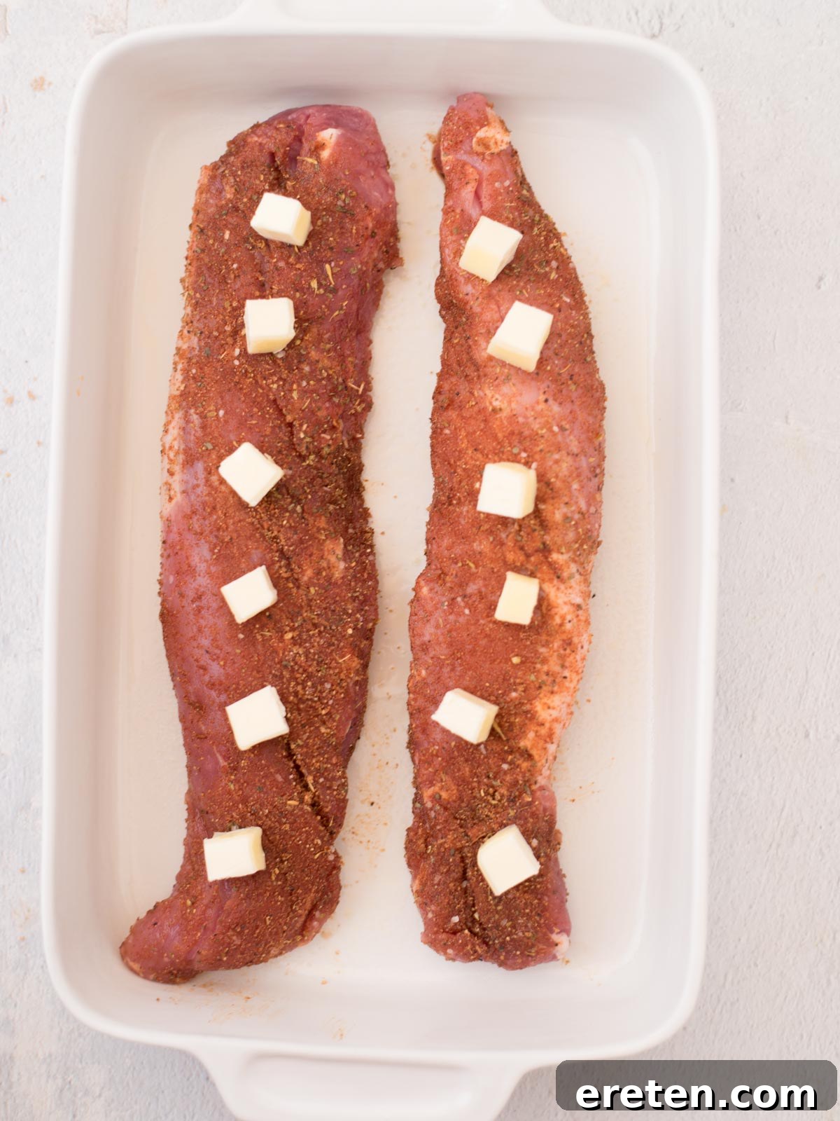 Raw pork tenderloin in a baking dish, seasoned and topped with small, evenly distributed cubes of butter, ready for roasting.