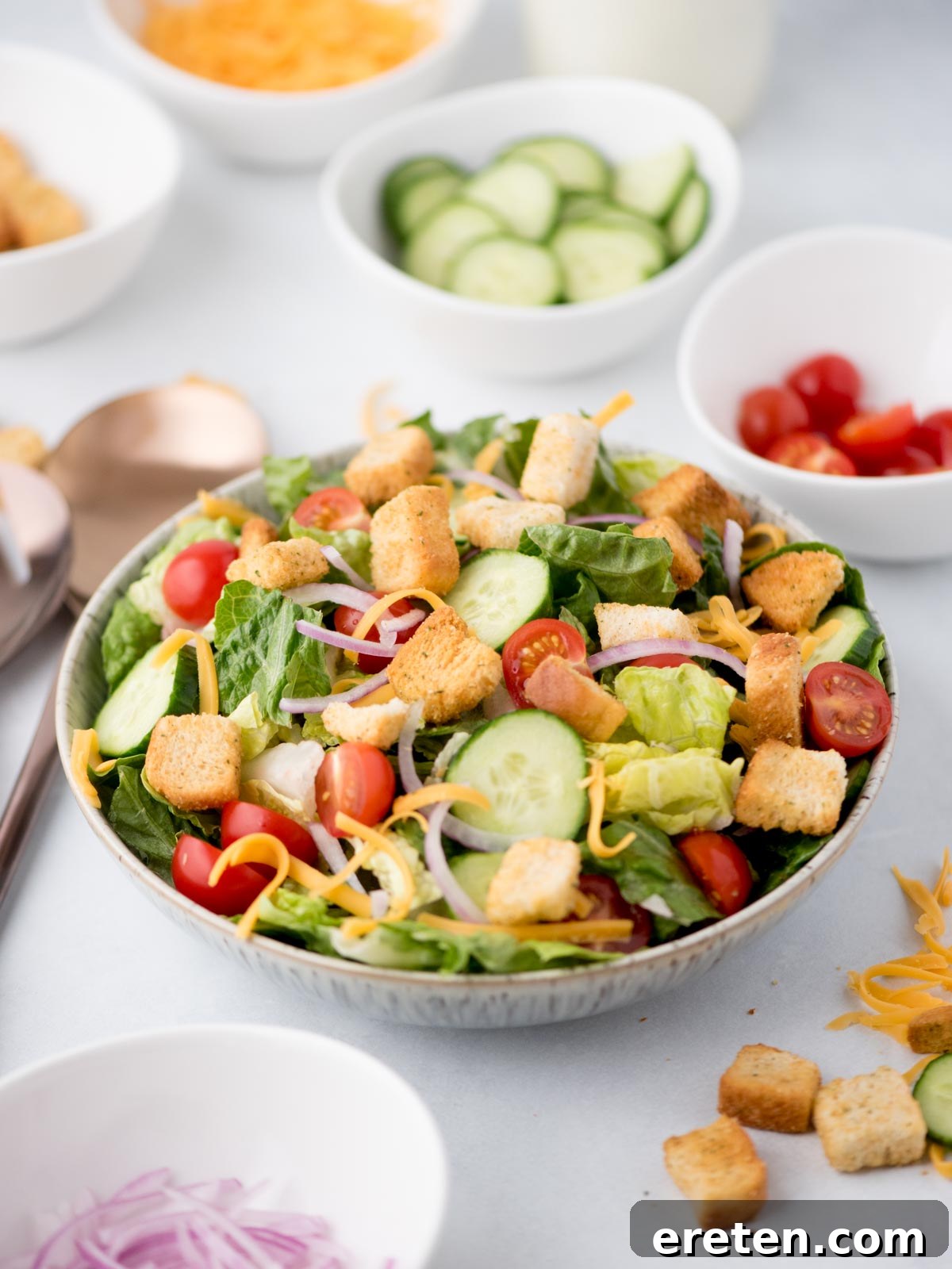 A vibrant house salad in a clear bowl, surrounded by fresh salad ingredients like cherry tomatoes, cucumber slices, and shredded cheese, with serving tongs ready to dish it out.