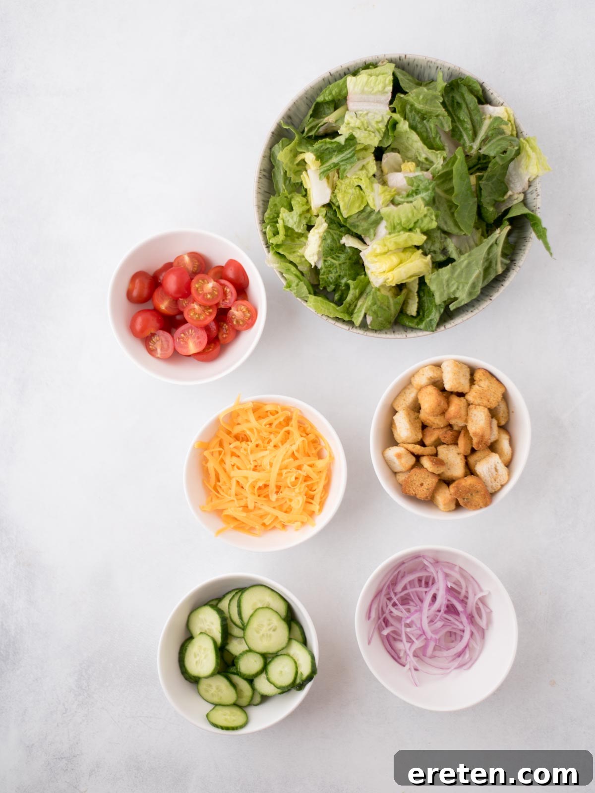 Individual ingredients for a house salad laid out on a white surface, including crisp romaine, colorful cherry tomatoes, a whole English cucumber, red onion slices, shredded cheddar cheese, and a pile of croutons.