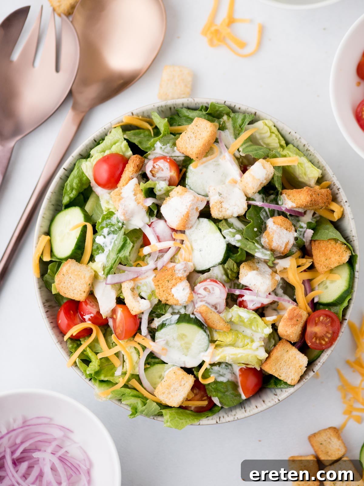 A beautifully presented house salad drizzled with creamy ranch dressing, with serving utensils alongside and a variety of fresh ingredients in the background, highlighting its readiness to serve.
