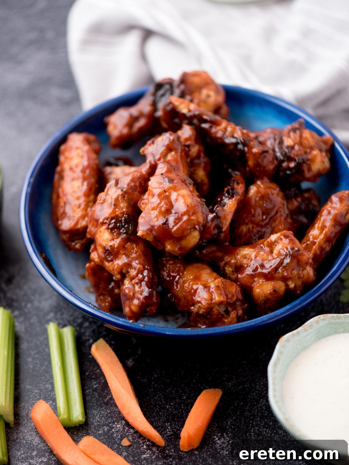 Succulent baked BBQ chicken wings, stacked high in a blue bowl, surrounded by a side of creamy ranch dip, crisp celery sticks, and vibrant carrot sticks for a perfect appetizer spread.