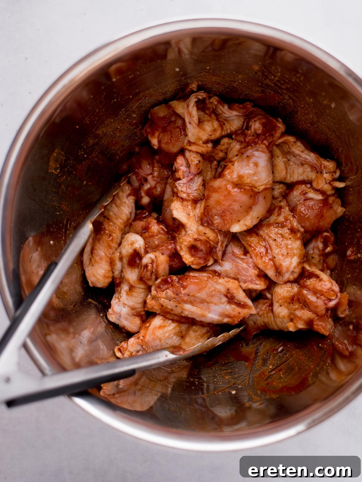 Uncooked chicken wings, fully coated in a vibrant mix of seasonings and thick barbecue sauce, nestled together in a stainless steel mixing bowl, ready for baking.