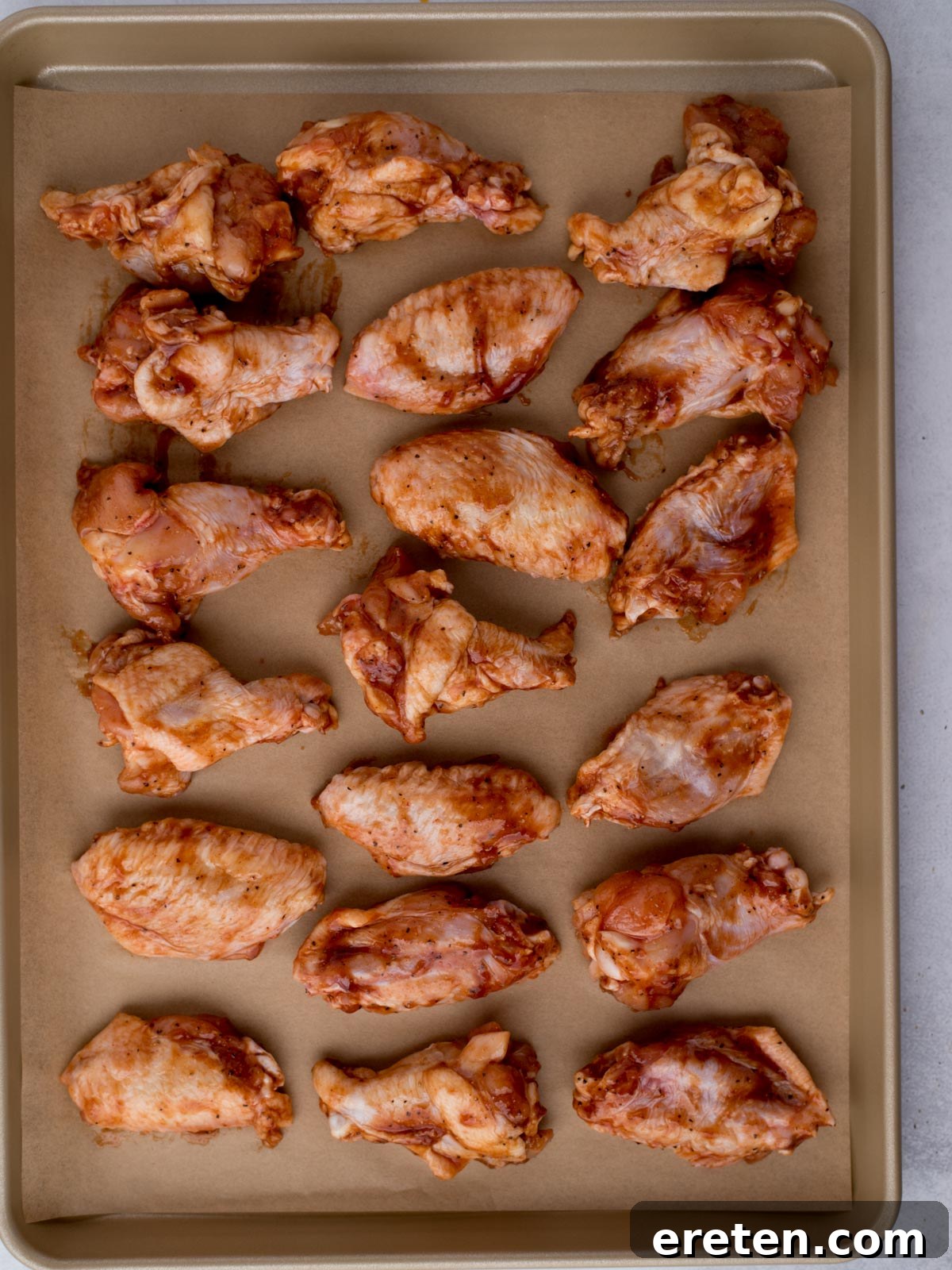 Uncooked chicken wings, perfectly coated in seasoning and rich barbecue sauce, neatly arranged in a single layer on a parchment-lined baking sheet, spaced for optimal crisping in the oven.