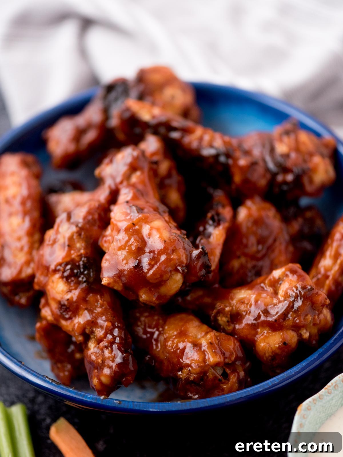 Beautifully baked BBQ chicken wings stacked high in a blue bowl, garnished with a sprinkle of fresh parsley, surrounded by bowls of creamy ranch and blue cheese dips, alongside refreshing celery and carrot sticks, creating an inviting and complete appetizer presentation.