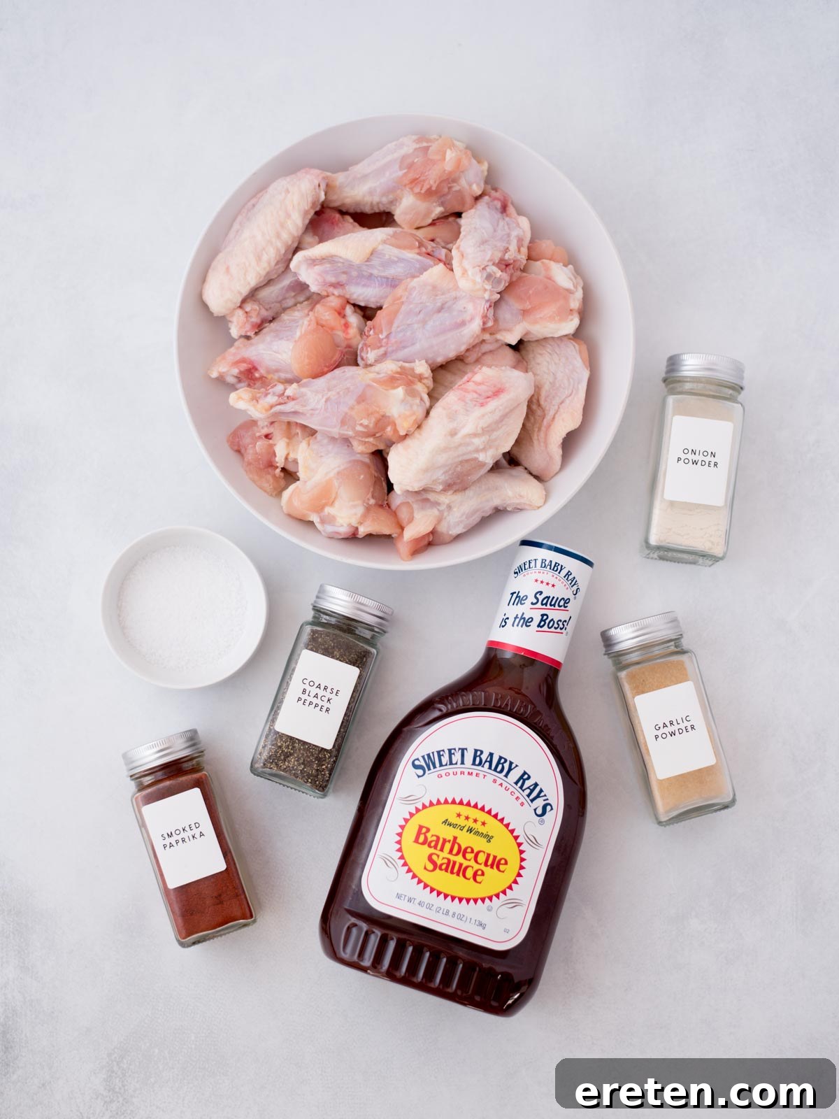 A vibrant selection of fresh ingredients laid out on a clean counter, including raw chicken wings, an array of spices like smoked paprika, garlic powder, onion powder, and coarse salt, along with a bottle of rich barbecue sauce, all ready for preparing delicious baked BBQ chicken wings.
