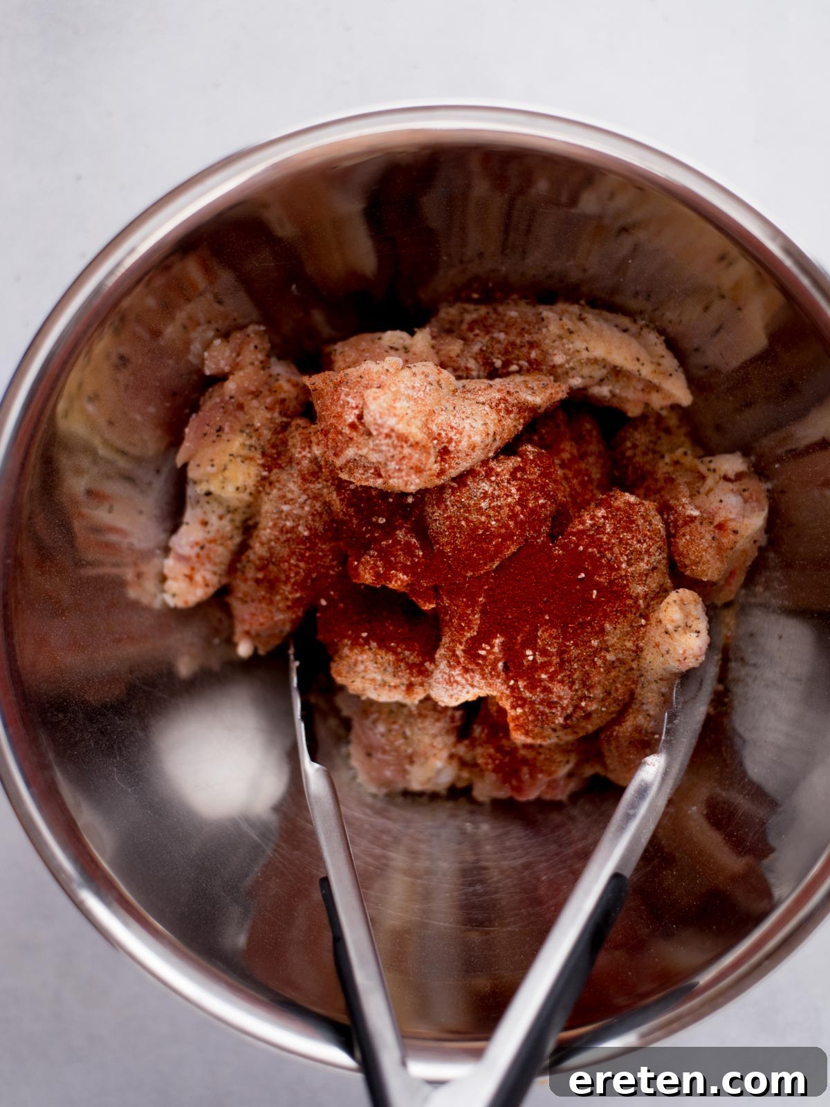 Raw chicken wings, seasoned with a flavorful spice rub, are ready for the oven in a shiny stainless steel mixing bowl, accompanied by a pair of metal tongs for easy handling.